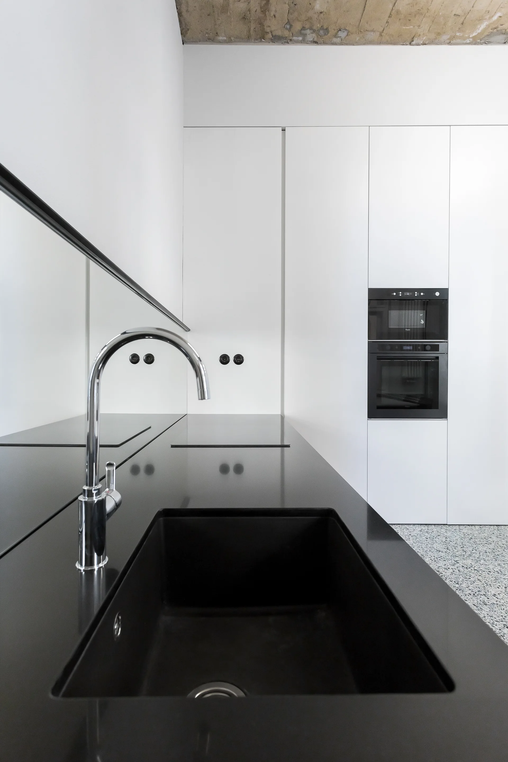 A modern minimalist kitchen with black stone countertops, an undermount sink, built-in double oven, and custom handleless white cabinetry.
