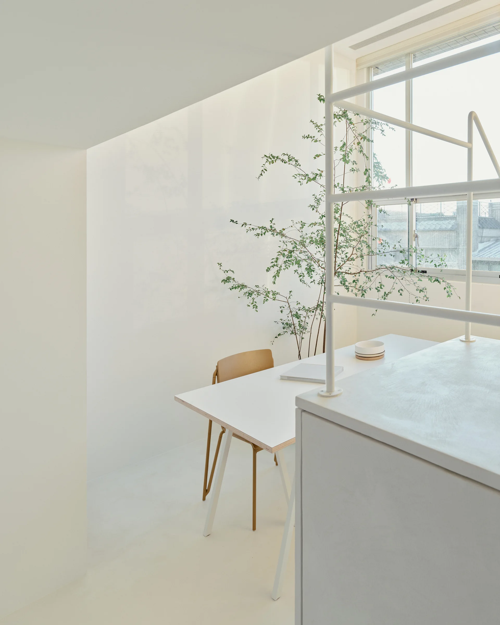 A minimalist white desk with a wooden chair sits in an all-white home office next to a tall green potted plant and a metal railing.
