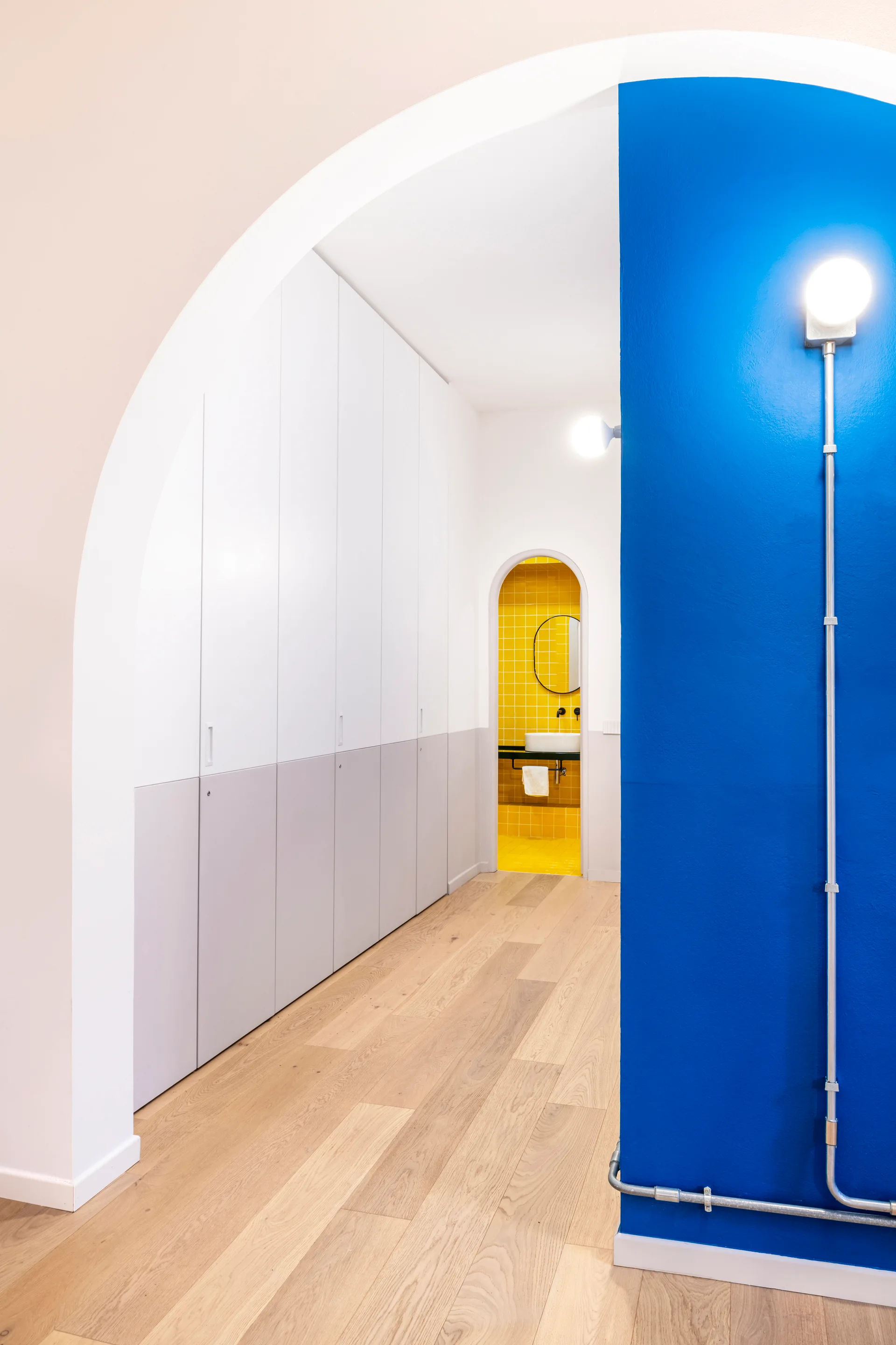 A hallway with light wood floors and two-tone built-in closets leading to a yellow tiled bathroom seen through an archway.