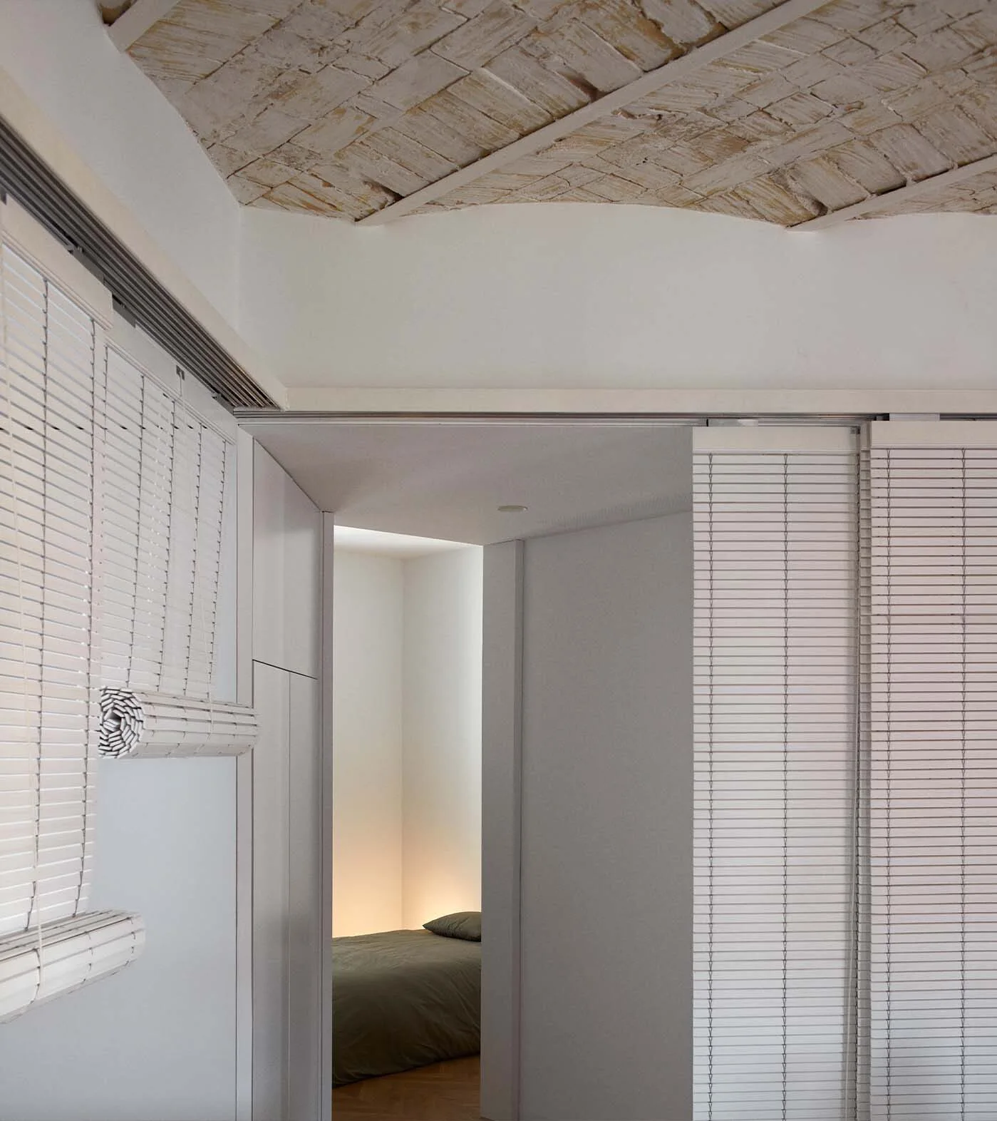 A view through a white doorway reveals a bed with muted green linens, set beneath an exposed white arched brick ceiling and flanked by white sliding blinds.