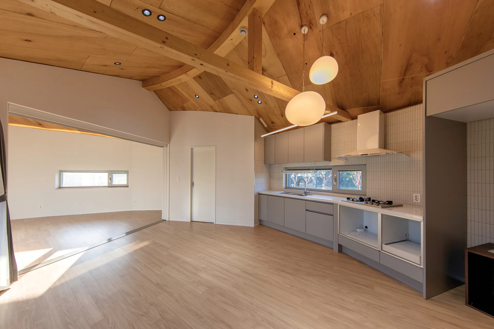 A minimalist kitchen features light grey cabinetry, vertical tile backsplash, two round pendant lights, and a vaulted wood-paneled ceiling with exposed beams.