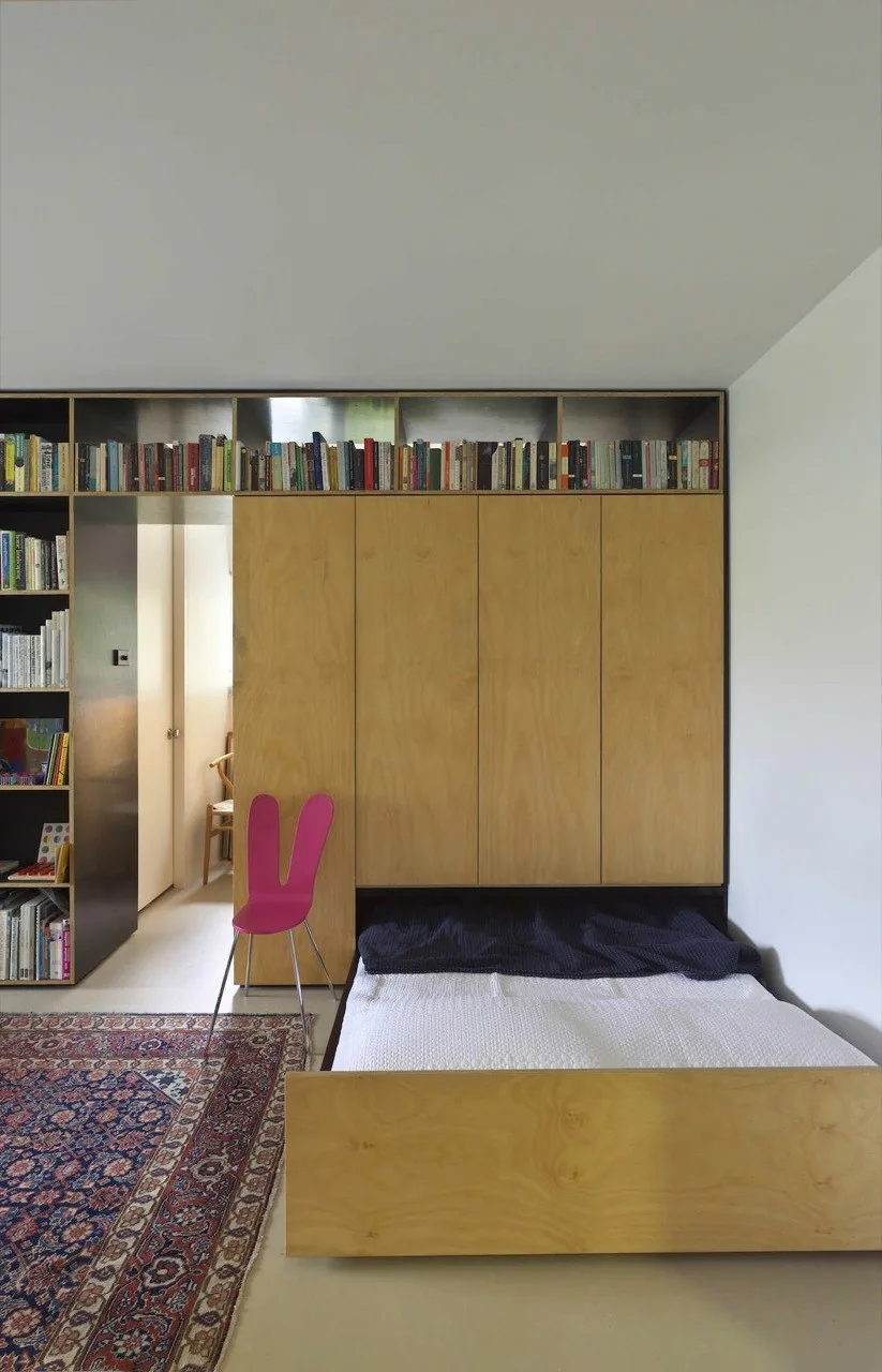 A light plywood wall unit with built-in storage and upper bookshelves stands behind a low platform bed next to a pink bunny ear chair.