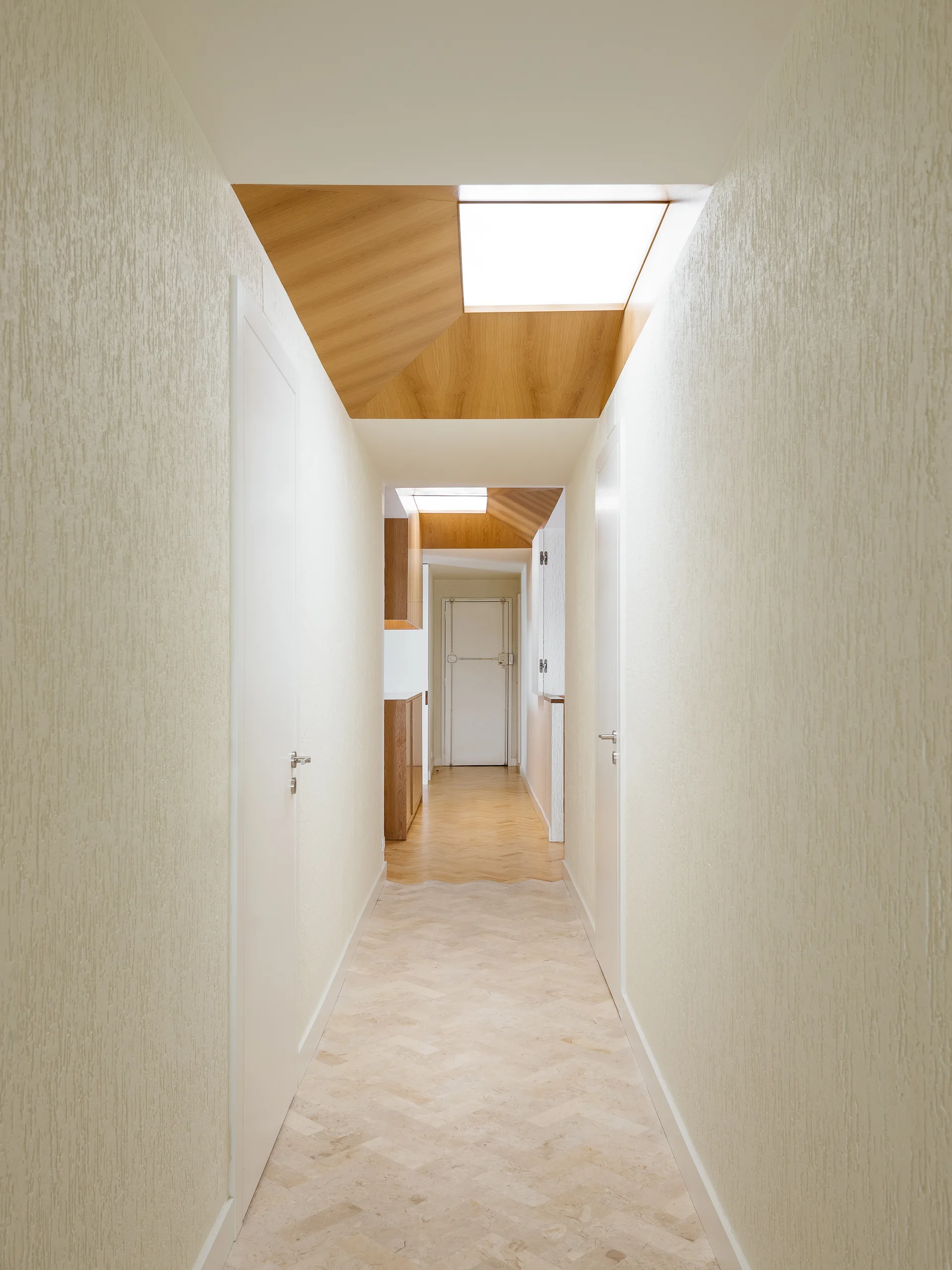 A long, narrow hallway features textured white walls, a light herringbone floor, and recessed wood-lined ceiling skylights.