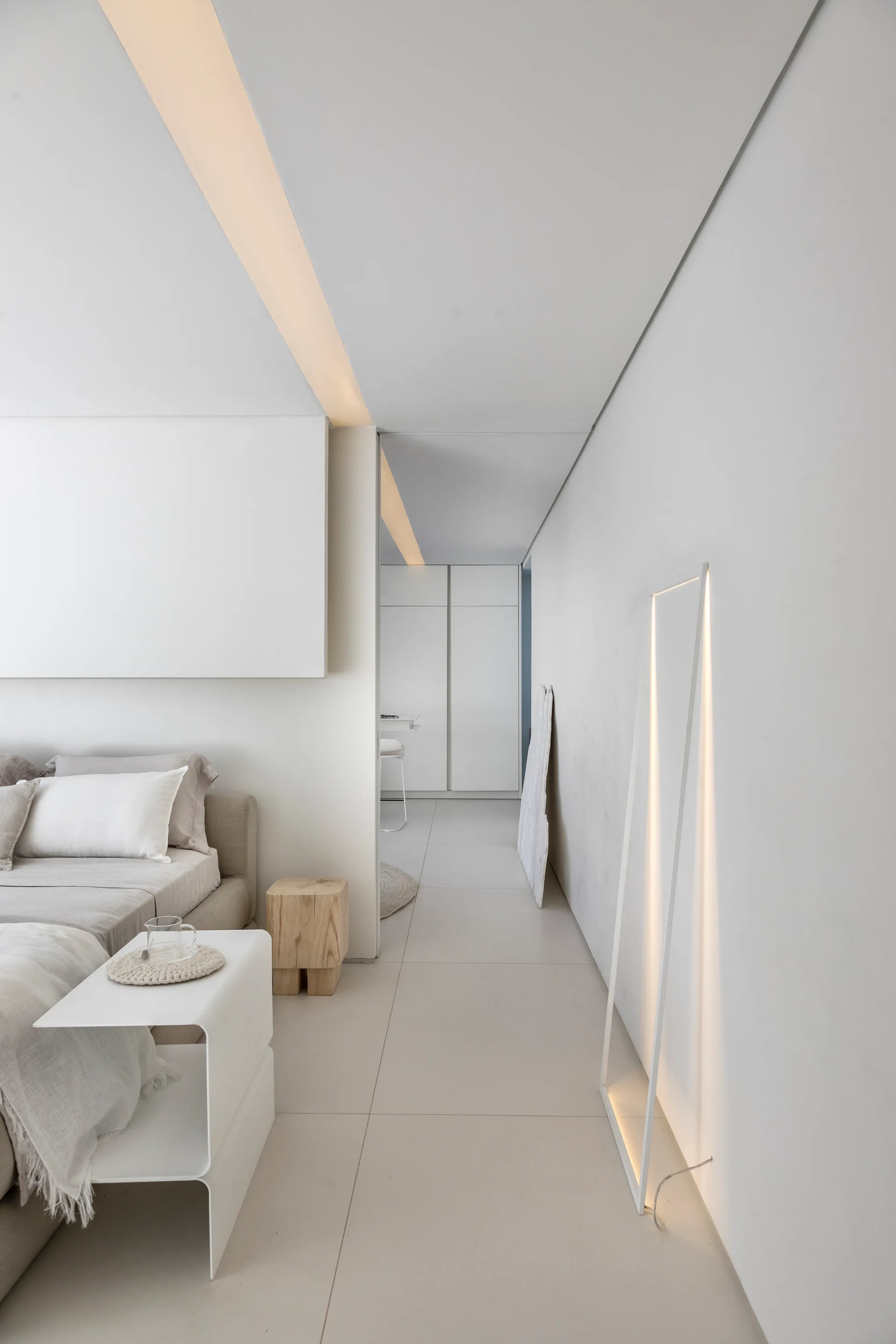 A minimalist white bedroom featuring a low-profile bed, curved metal nightstand, wood block stool, and recessed linear ceiling lighting.