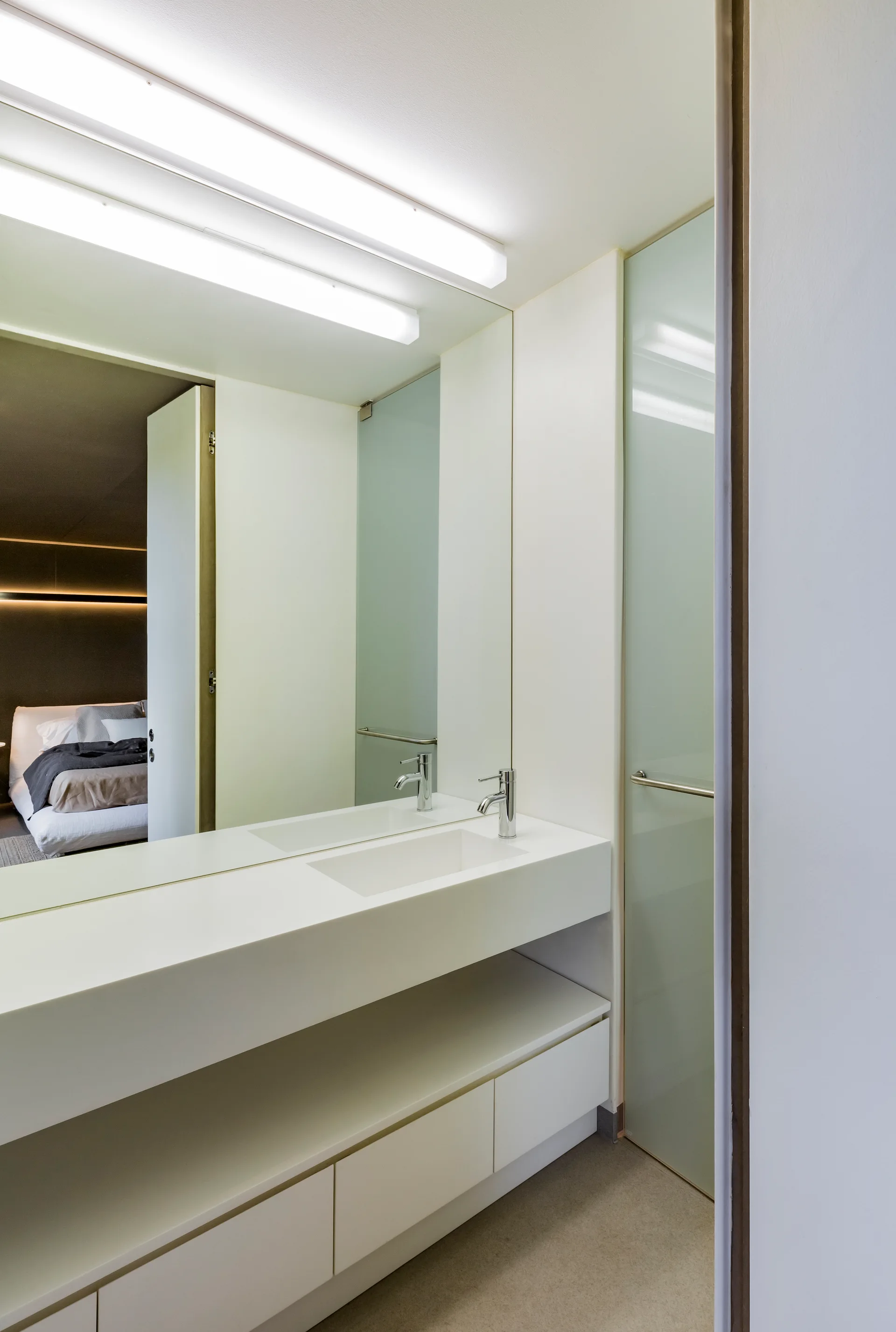 A minimalist white bathroom features a floating vanity with an integrated sink, a large wall mirror, and a frosted glass door.