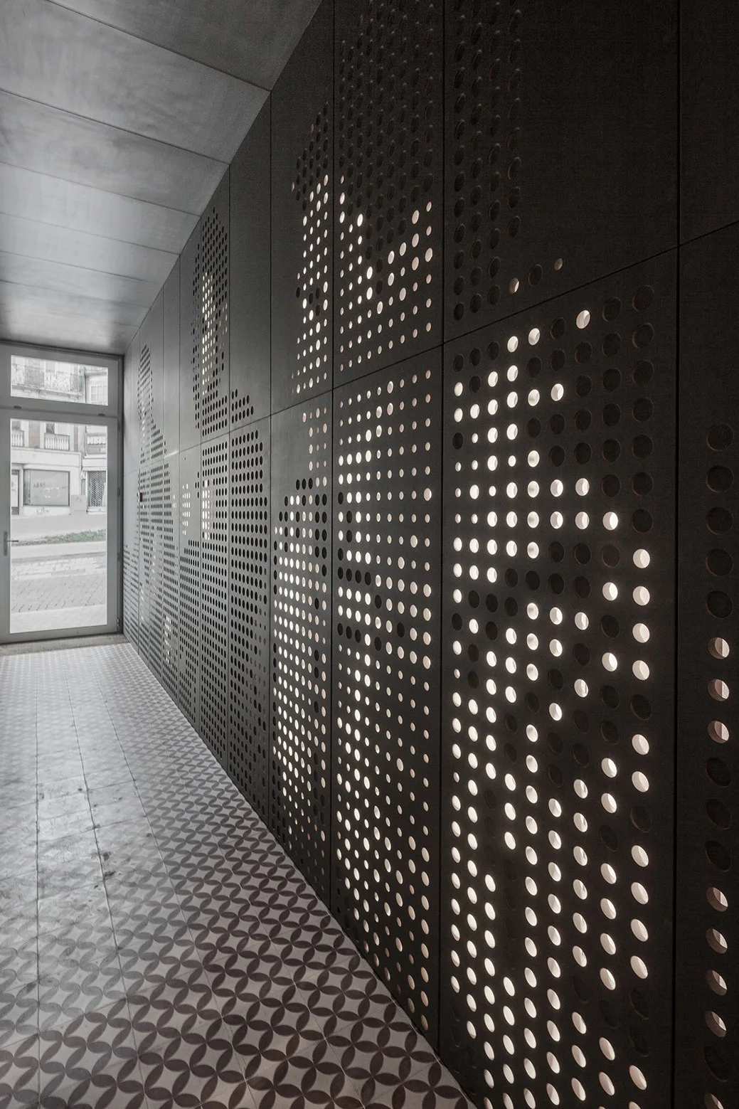 A long hallway features dark perforated metal wall panels with warm integrated backlighting and a patterned geometric tile floor leading to a glass door.