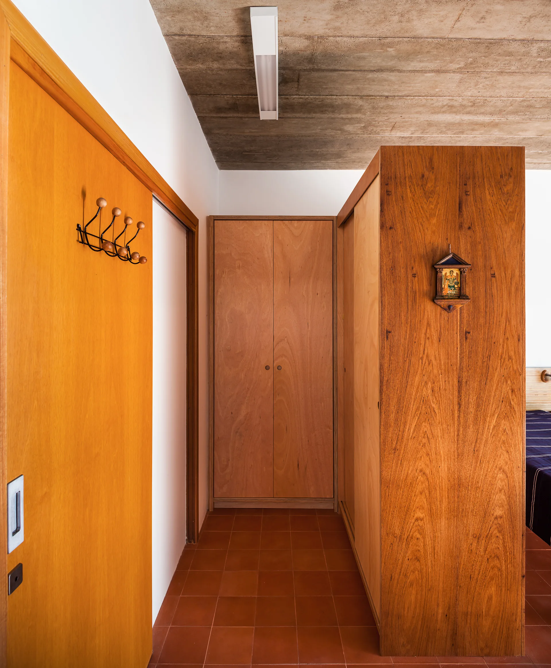 A modern hallway features built-in warm wood cabinetry, reddish-orange terracotta floor tiles, and an exposed concrete ceiling with a linear light fixture.
