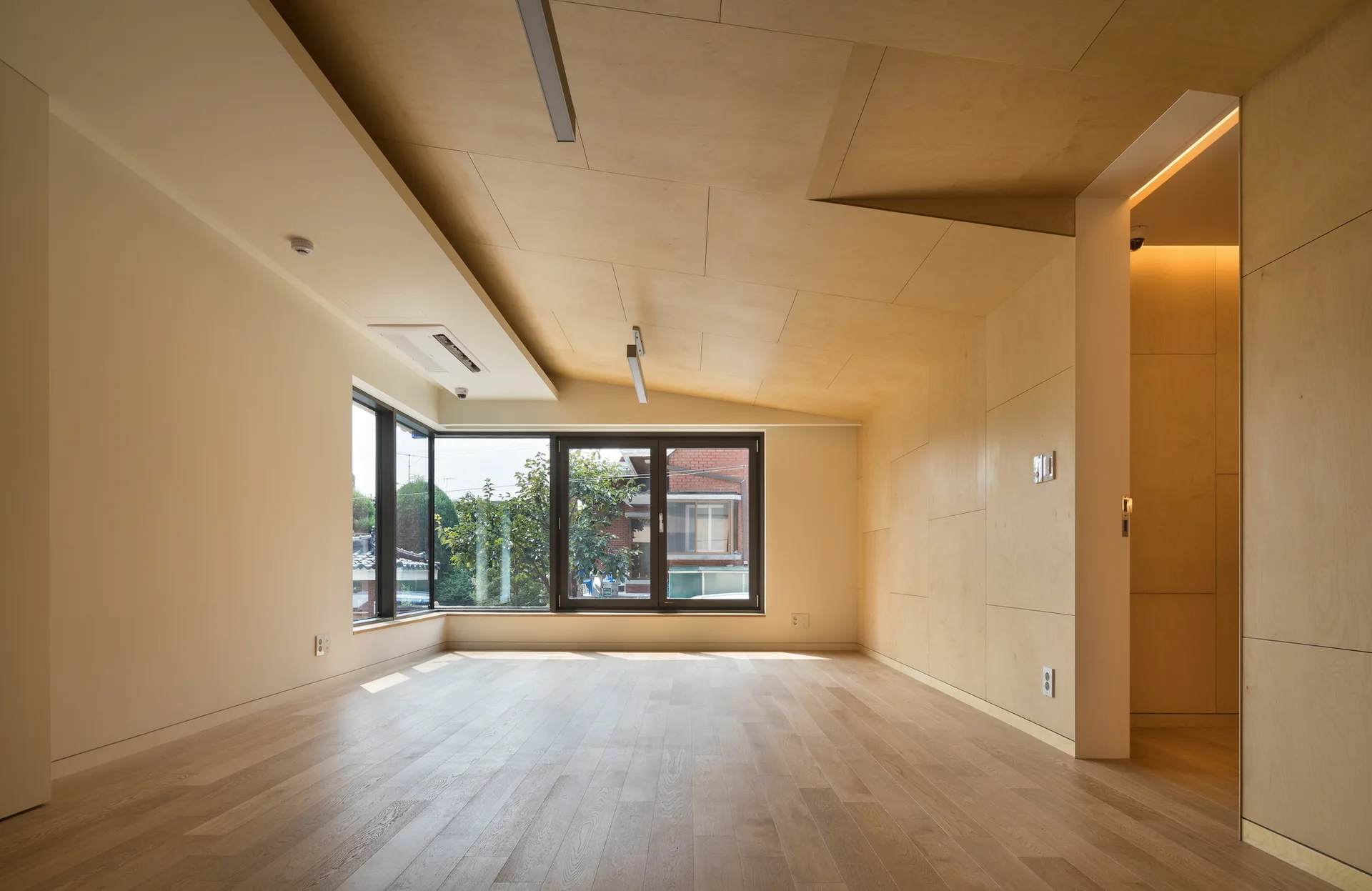 An empty room features light wood plank flooring, a sloped ceiling lined with plywood panels, and large black-framed windows.