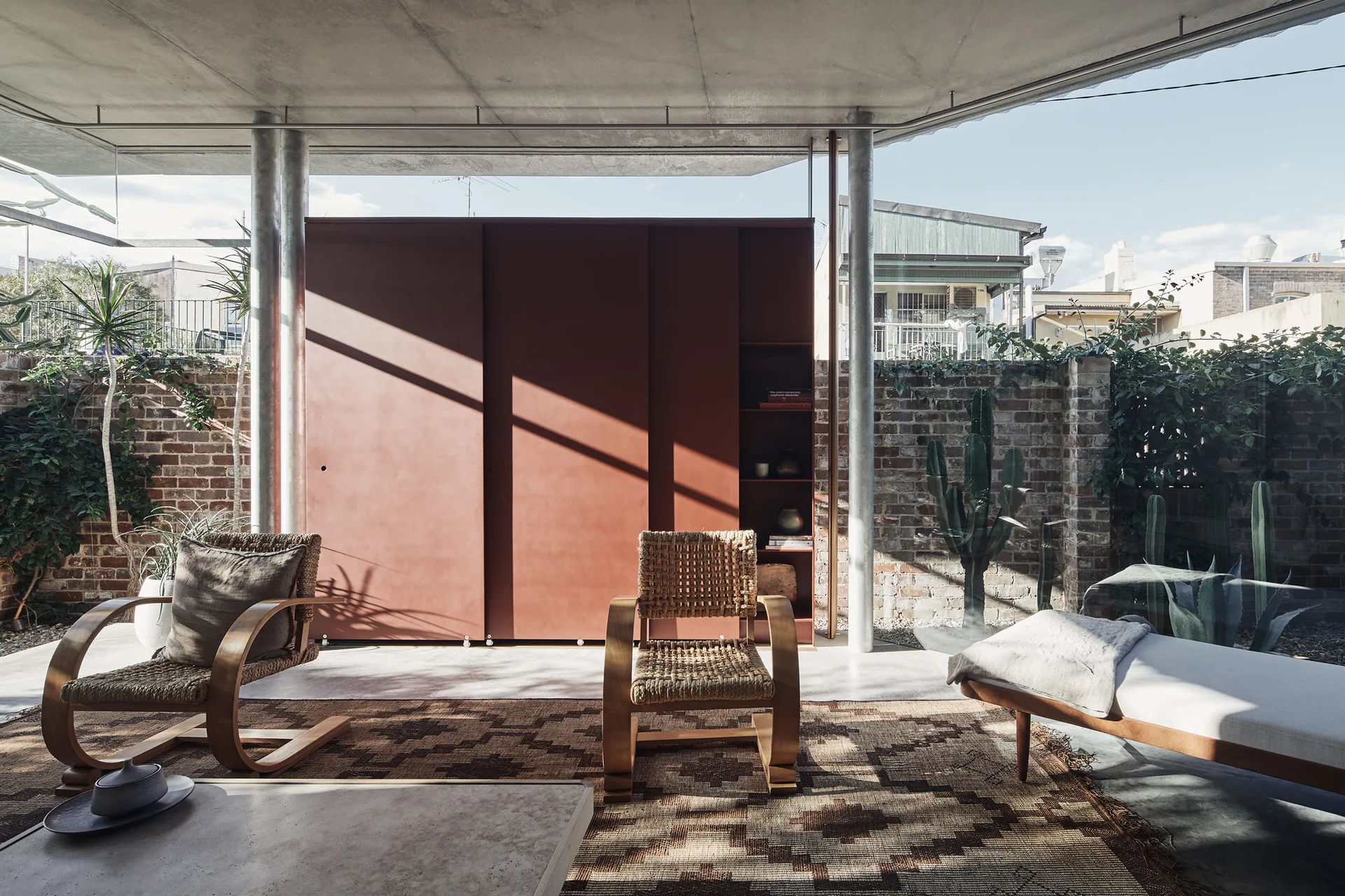 A covered concrete patio features a large terracotta sliding cabinet, mid-century woven armchairs on a patterned rug, and an exposed brick wall backdrop.