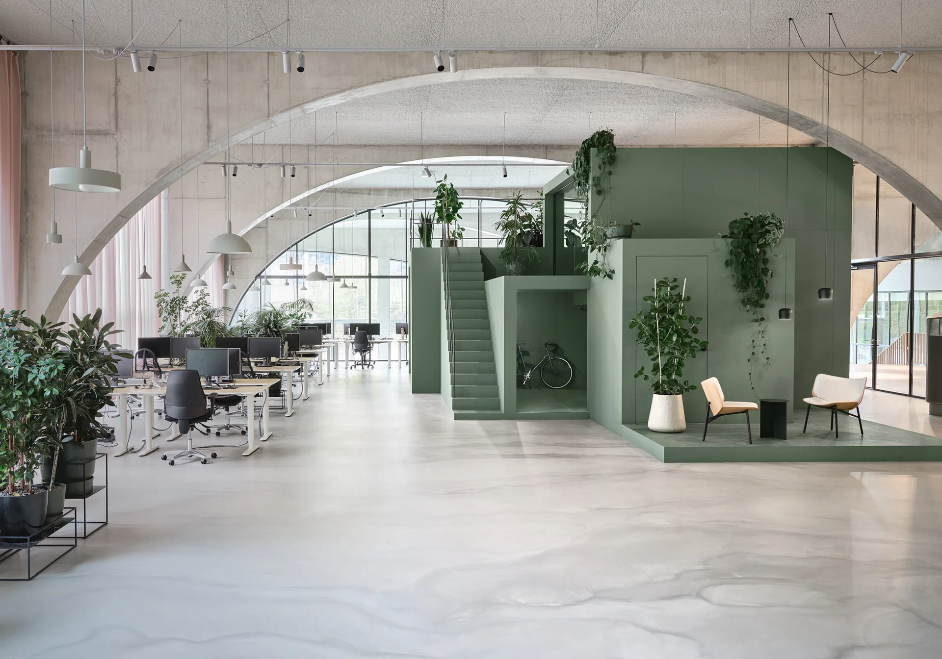 A spacious open-plan office features massive concrete arches, clusters of desks, and a multi-level sage green geometric structure with a staircase.