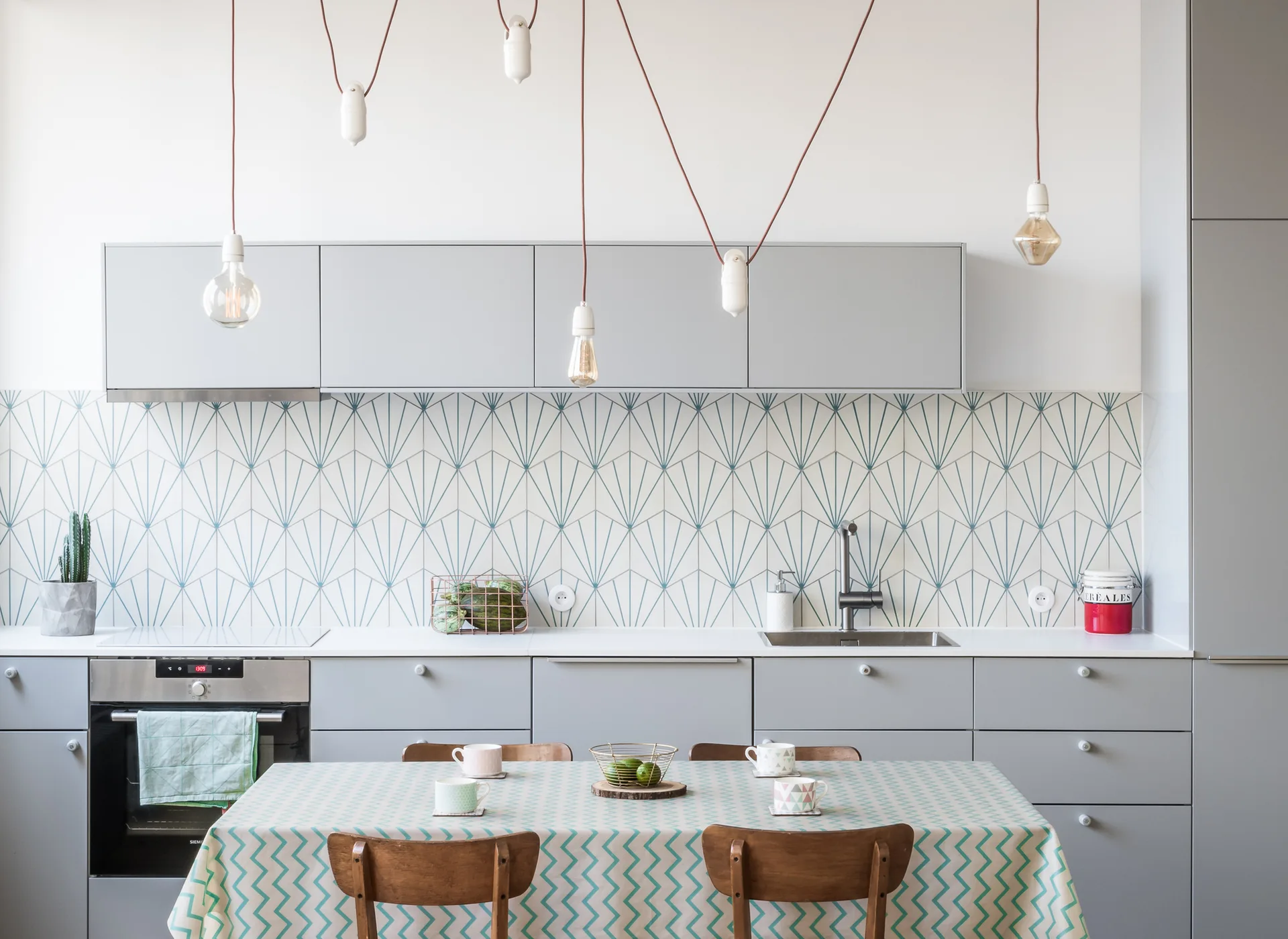 A light grey kitchen features a blue-and-white geometric tiled backsplash, flat-front cabinets, and a wood dining table set with a patterned tablecloth.