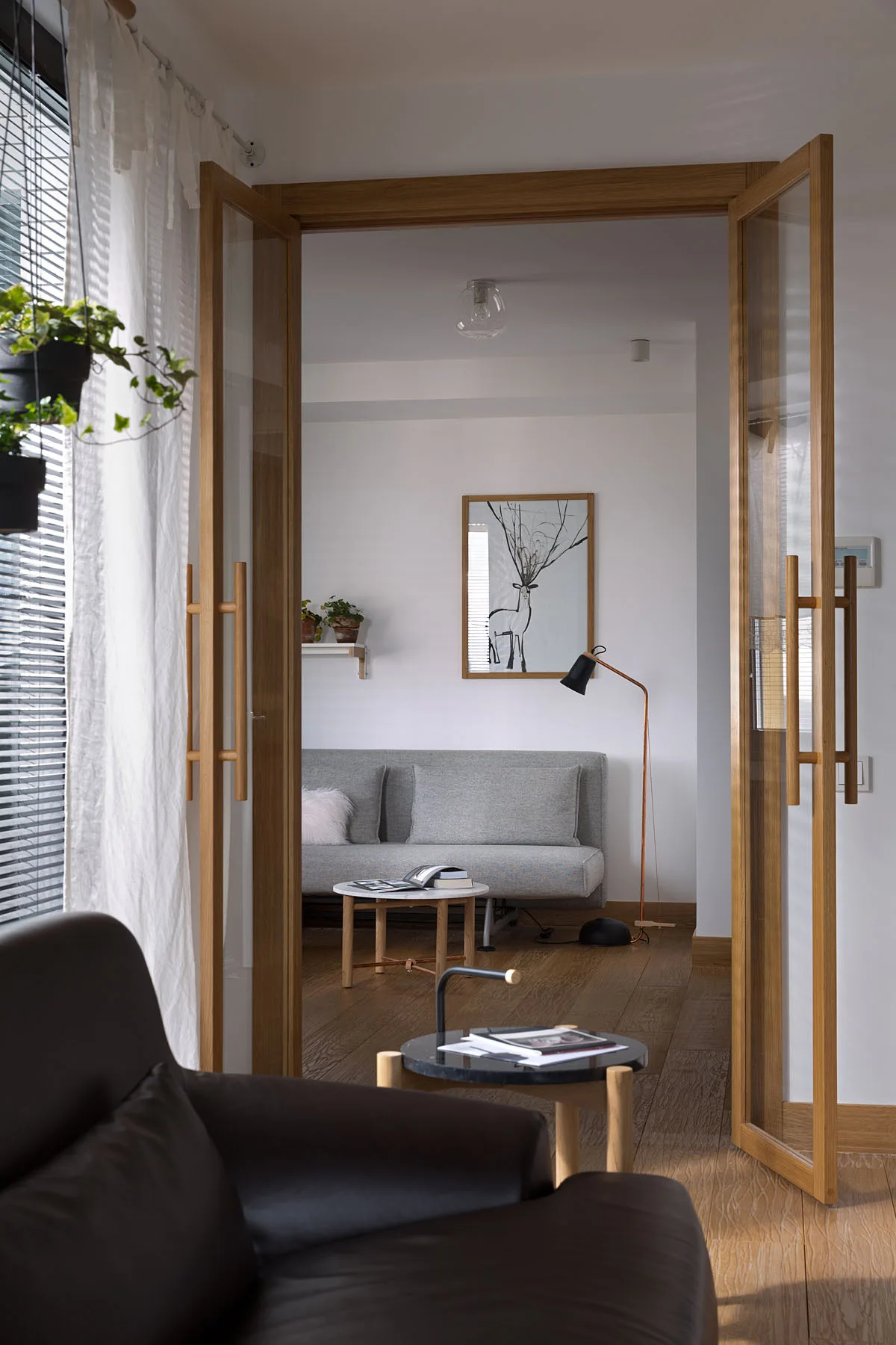 A view through open wooden-framed glass double doors into a minimalist living room with a gray sofa, wood flooring, and a slim floor lamp.