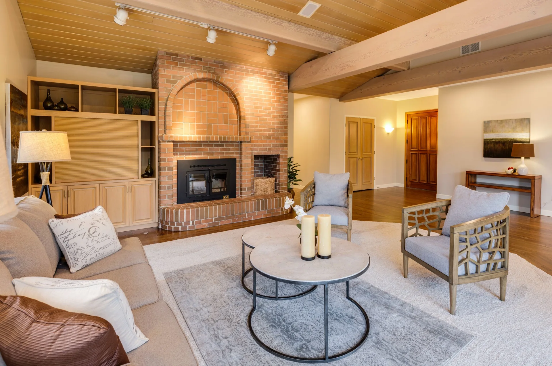 A living room features a large arched brick fireplace, exposed wood ceiling beams, a beige sofa, and two gray accent chairs around nested coffee tables.
