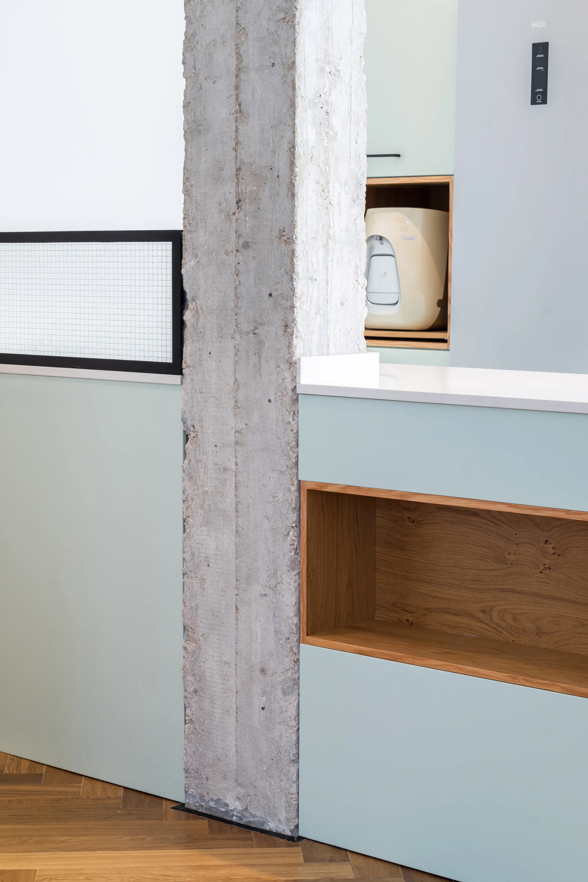 Sage green kitchen cabinetry built around a raw concrete pillar features natural oak wood shelf inserts and herringbone wood flooring.