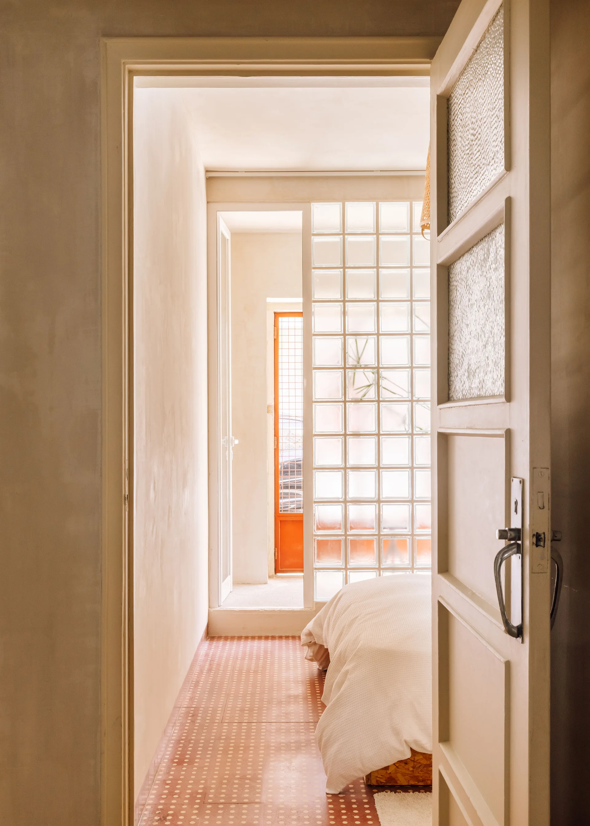 A view through an open doorway into a bedroom featuring a glass block partition wall, patterned terracotta floor tiles, and a bed with white linens.