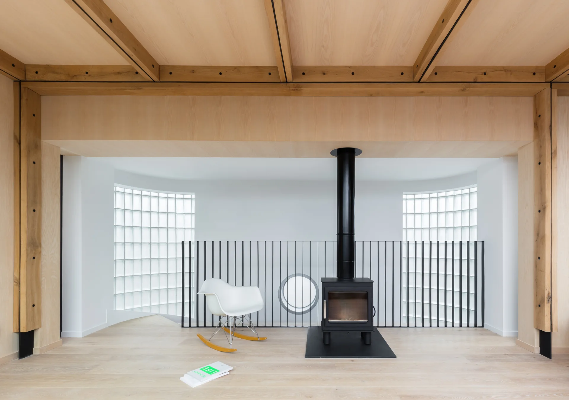 A minimalist interior room featuring a light wood ceiling, a black wood-burning stove, curved glass block windows, and a white rocking chair.