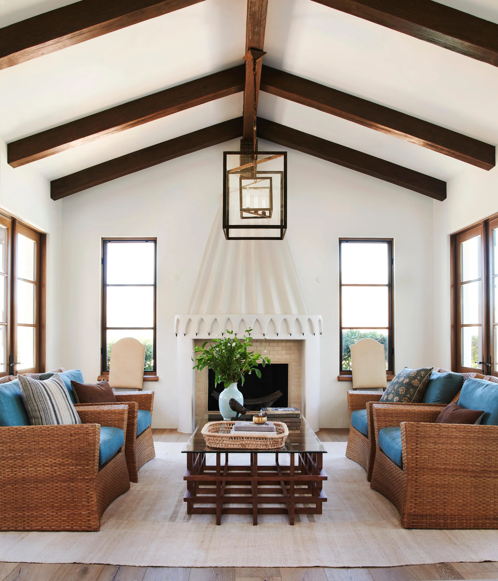 A symmetrical living room with four blue-cushioned wicker chairs around a glass coffee table, a central white plaster fireplace, and dark ceiling beams.