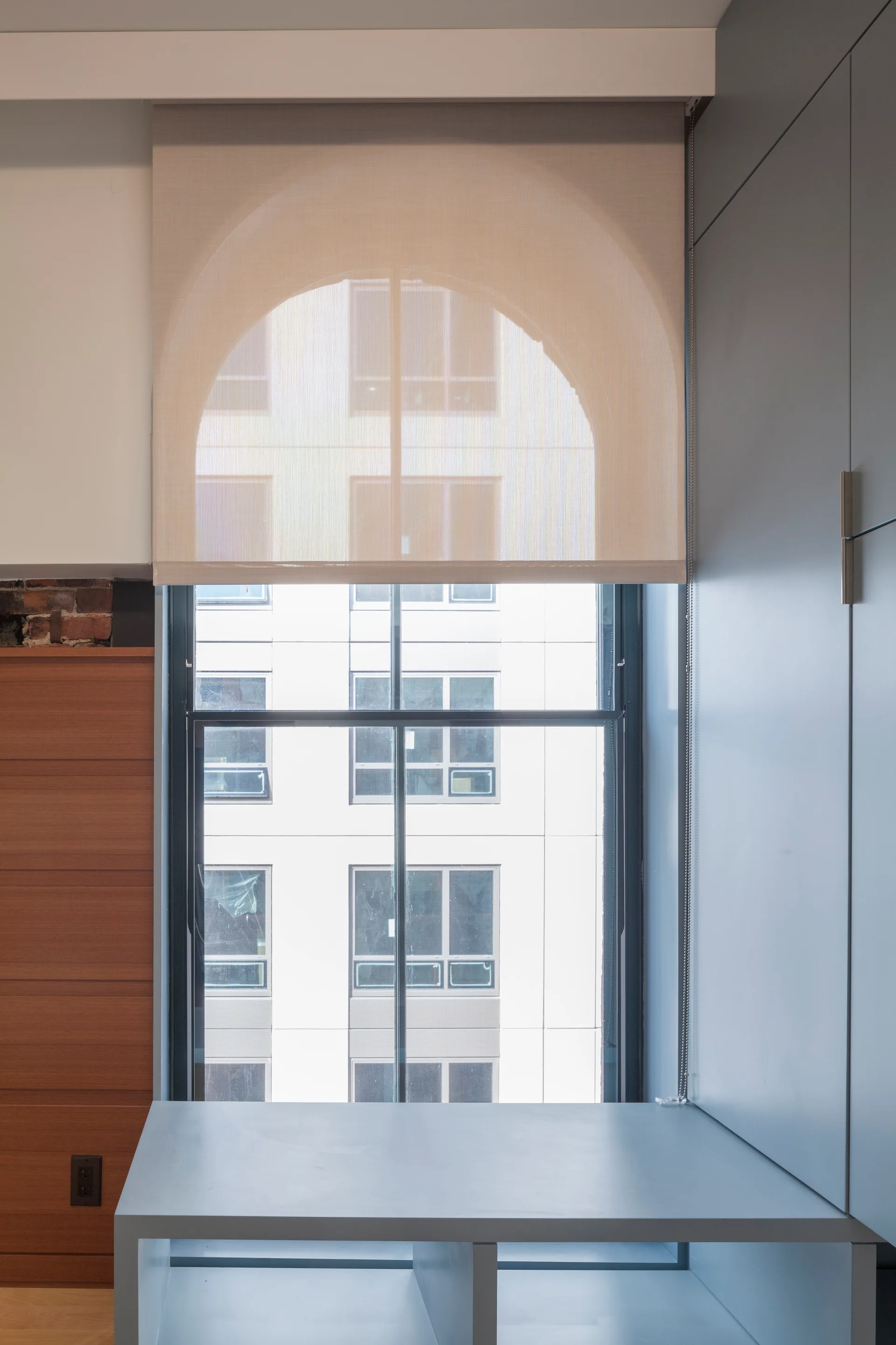 A modern window with a semi-sheer arched roller shade is nestled between grey built-in cabinetry and warm wood paneling.