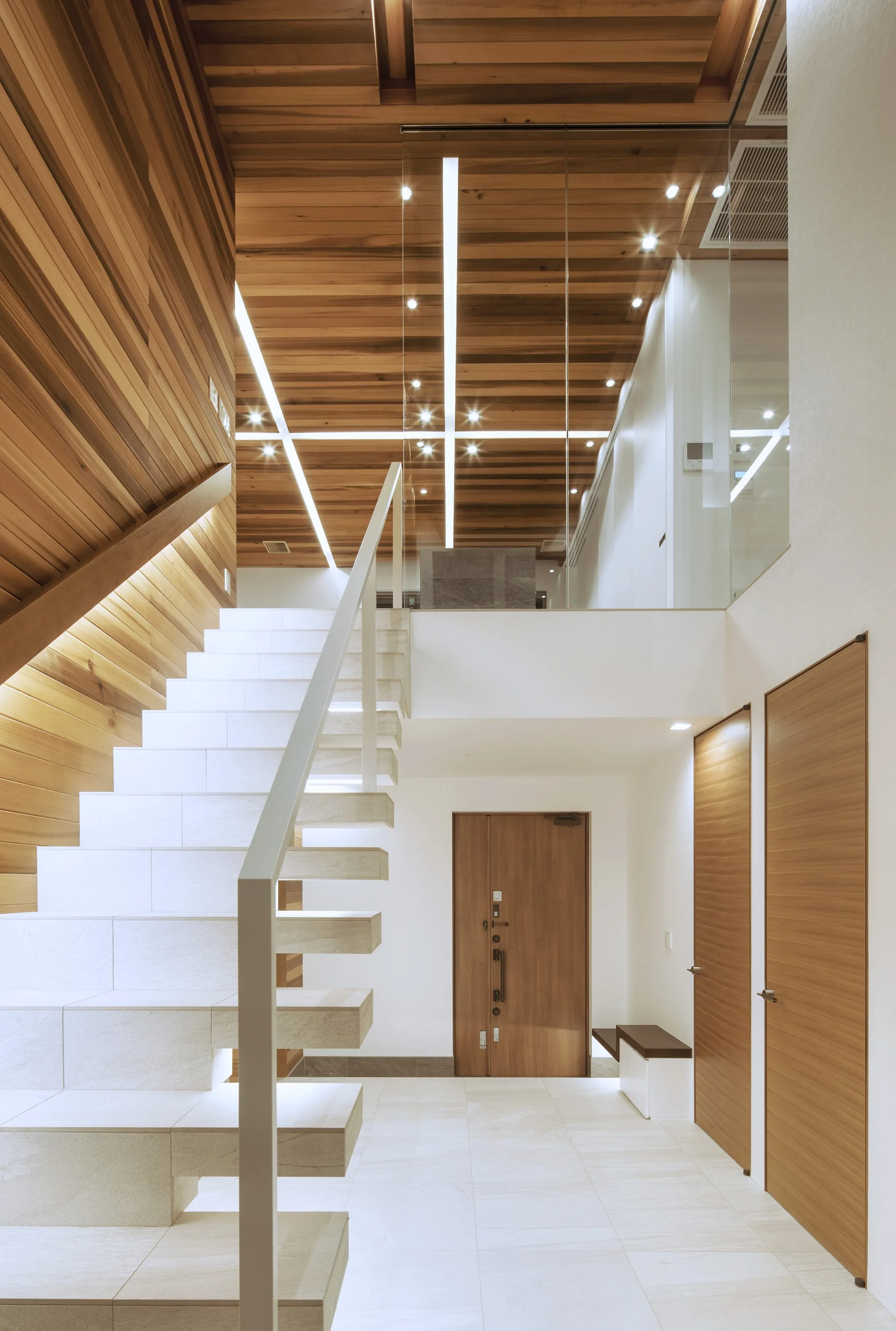 A modern entryway features a floating stone staircase, warm wood-paneled walls and ceiling, and a geometric pattern of recessed linear lights.