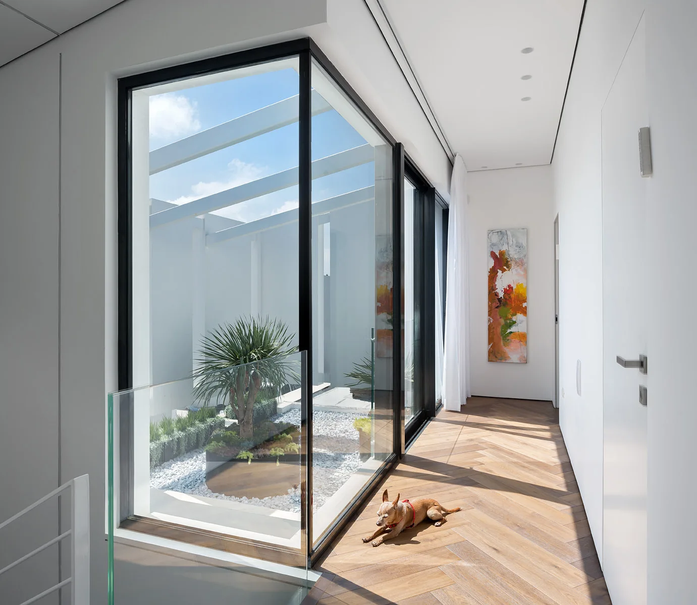 A bright modern hallway features herringbone patterned wood flooring and a large black-framed window looking into a central interior atrium.