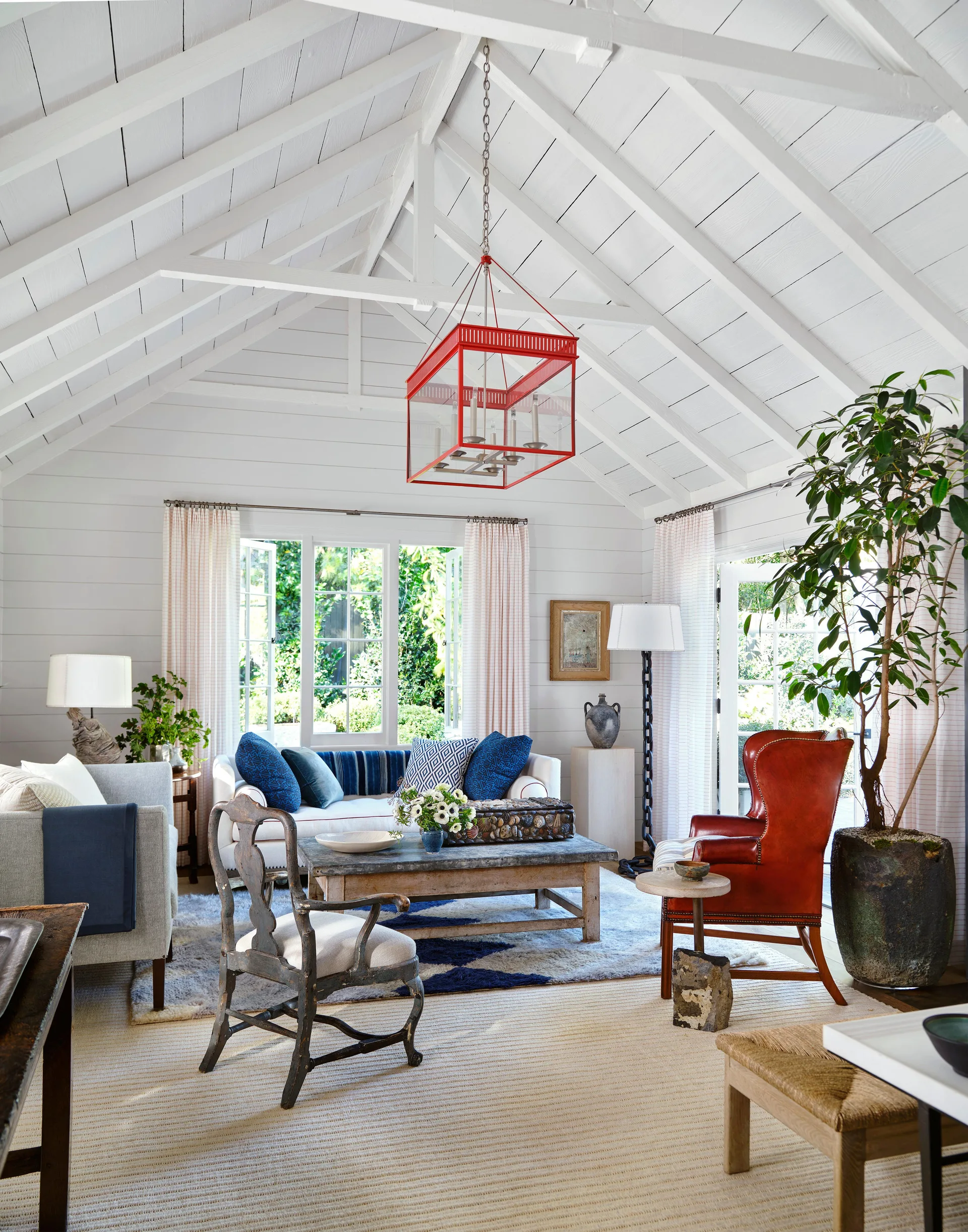 A sunlit living room featuring a vaulted white wood ceiling, a red lantern pendant light, a white sofa with blue pillows, and a leather wingback chair.