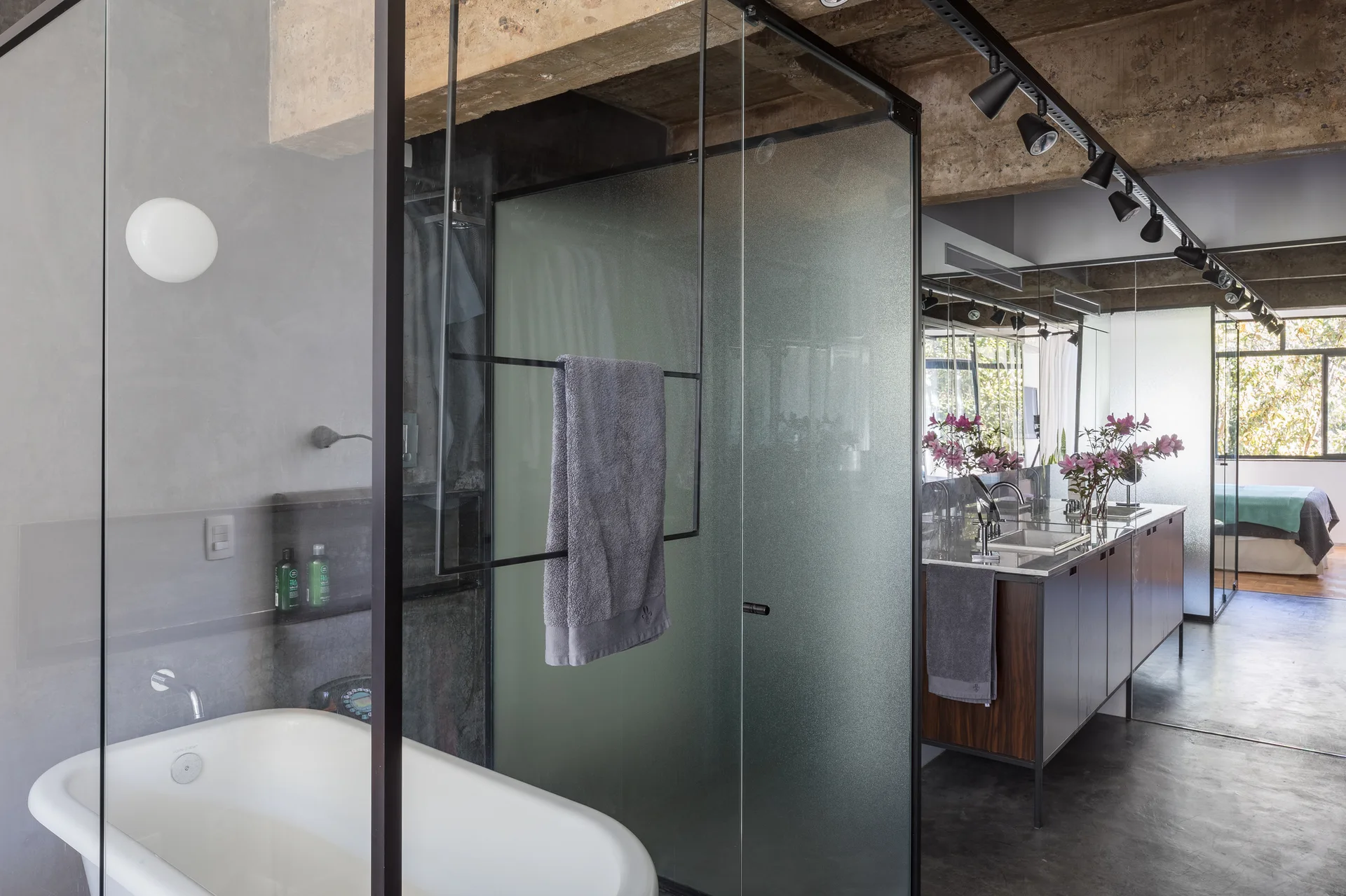 A modern industrial bathroom featuring a white freestanding bathtub next to a black-framed frosted glass shower enclosure under an exposed concrete ceiling.