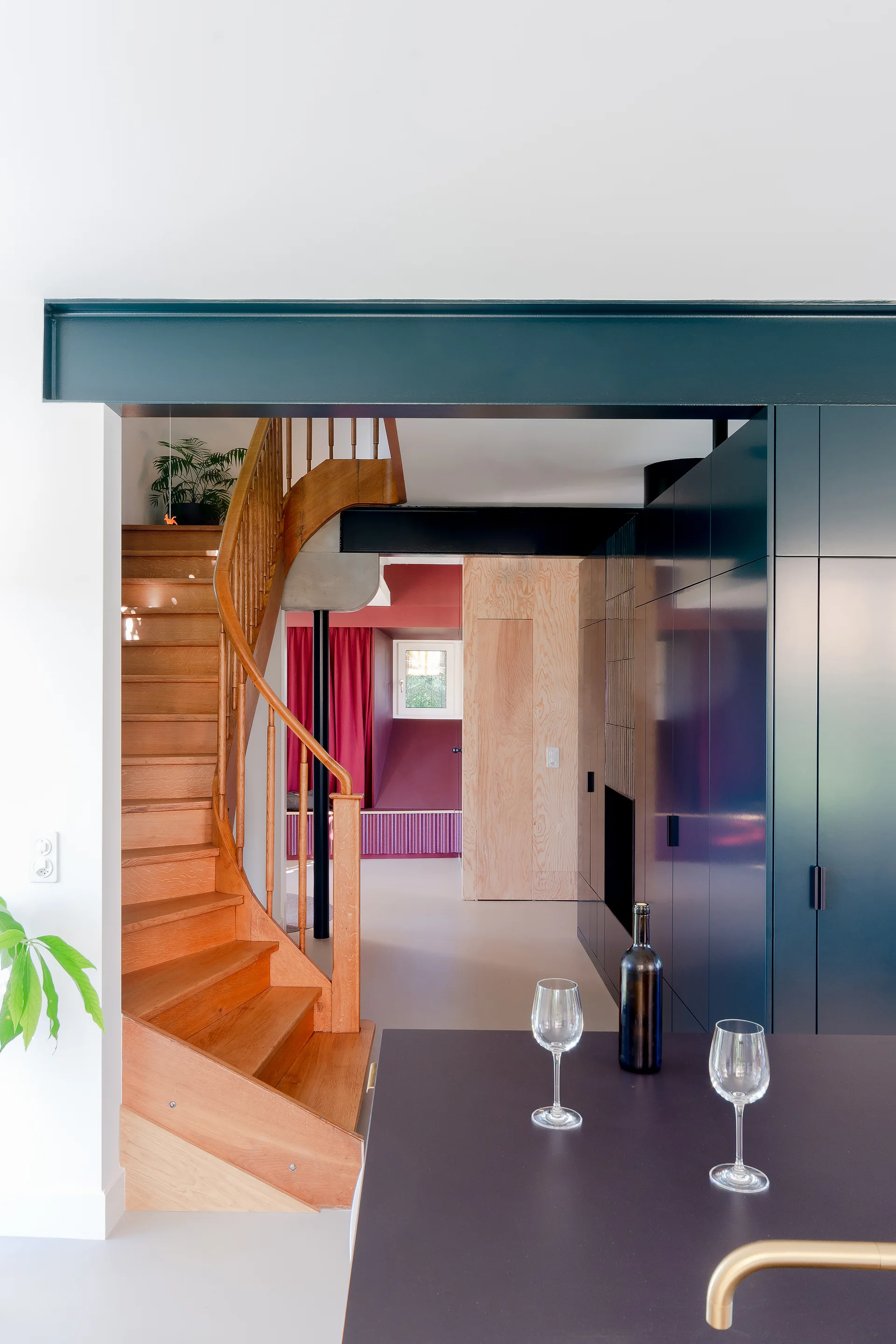 A modern interior looking through to a curved wooden staircase next to deep blue built-in cabinets under a dark teal structural beam.