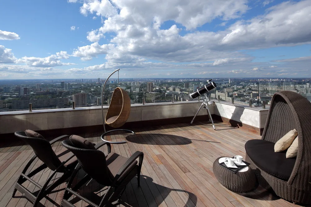 A rooftop terrace with dark brown wicker lounge chairs, a hanging egg chair, and a telescope overlooking a city skyline under a blue sky.