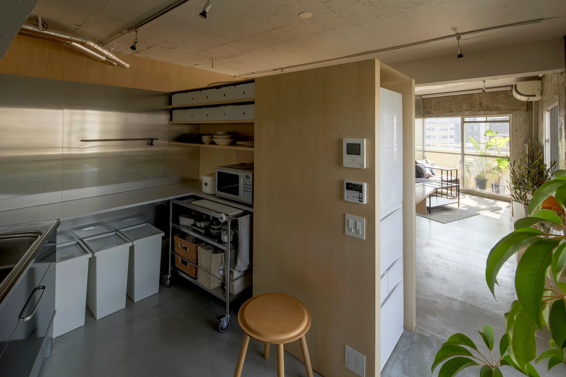 A minimalist kitchen features a light plywood room divider, stainless steel countertops, and matching white trash bins beneath an exposed concrete ceiling.