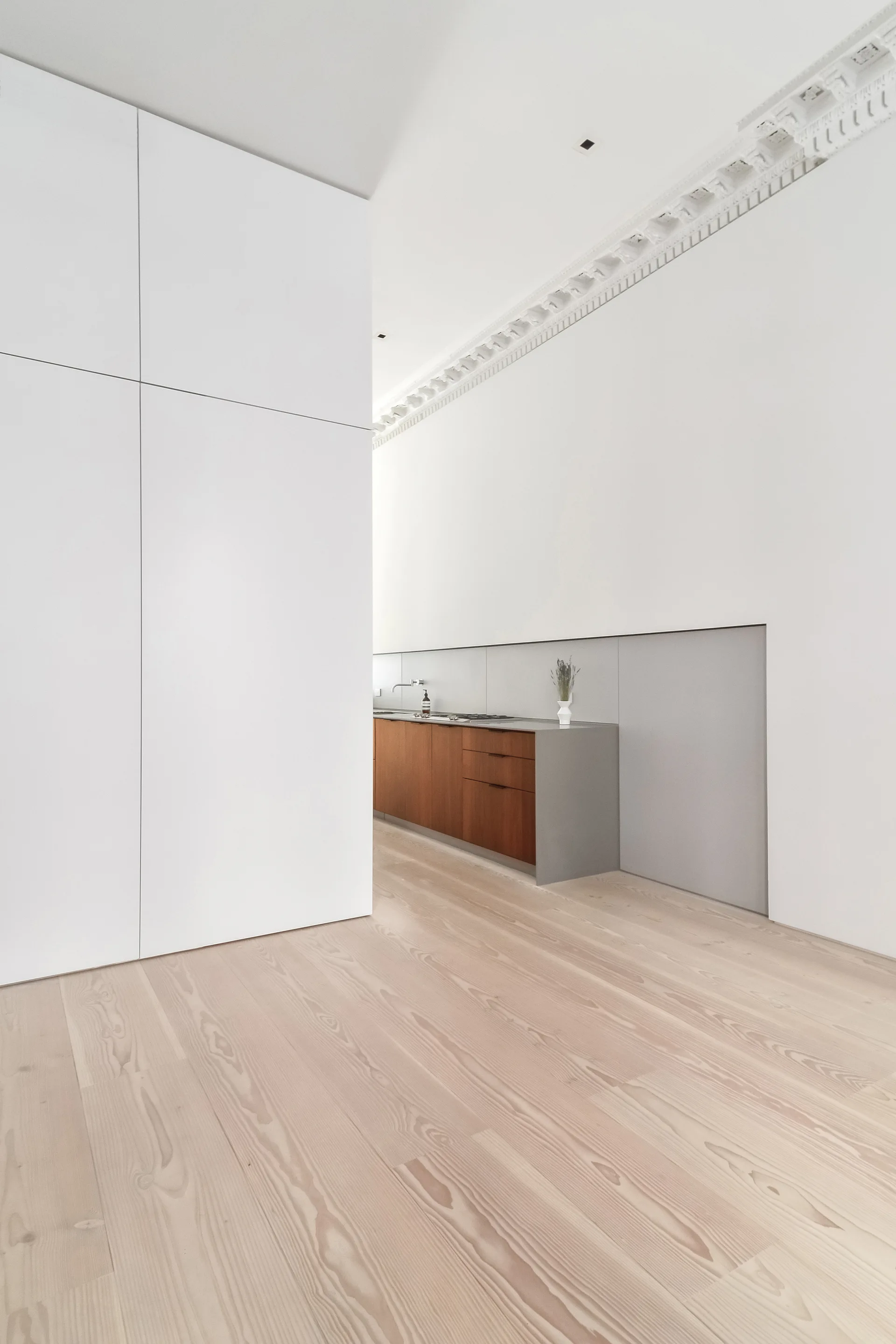 A minimalist kitchen features natural wood base cabinets and a grey countertop set within a white wall panel alcove beneath ornate crown molding.