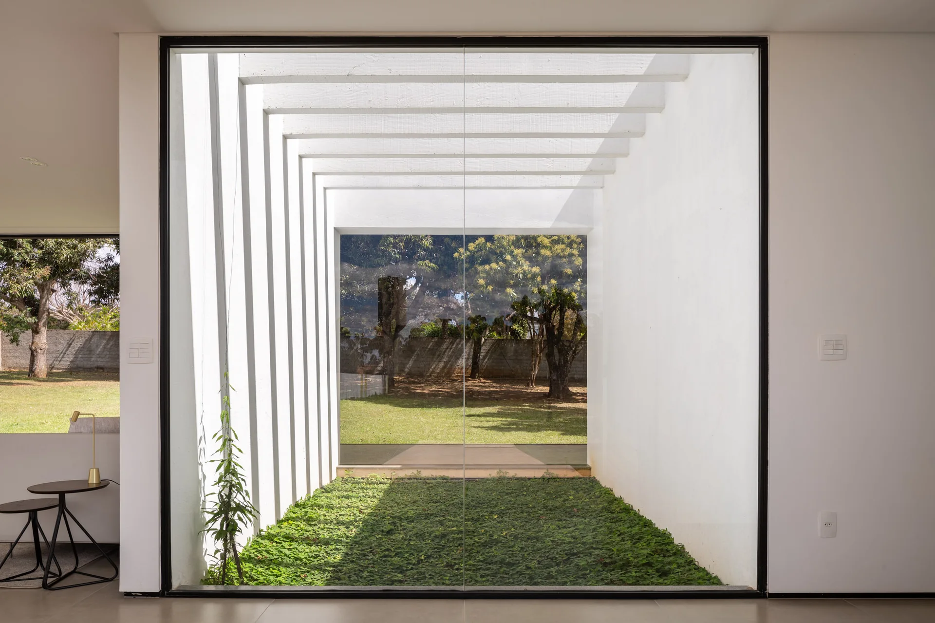 A minimalist corridor features a floor-to-ceiling glass wall framing an indoor courtyard with green groundcover under white overhead beams.