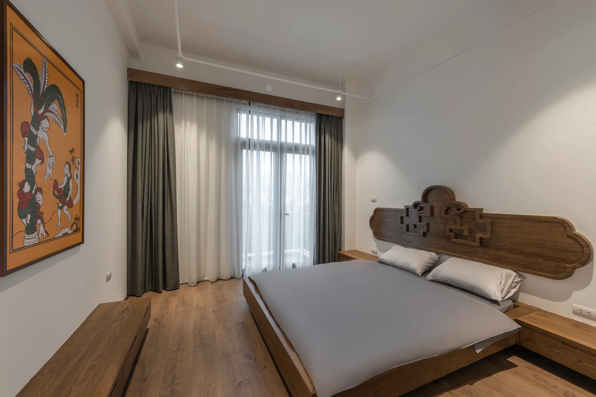 A minimalist bedroom features a wooden platform bed with a large carved headboard, matching low bedside tables, white walls, and wood flooring.