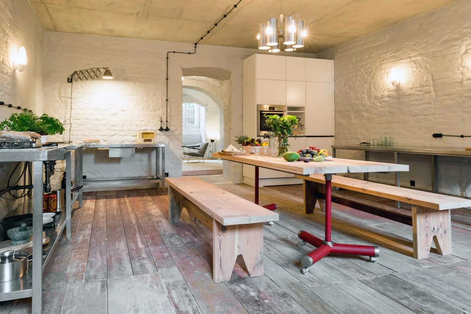 A rustic kitchen and dining space features white painted brick walls, a long wooden table with red metal legs, wood benches, and weathered floorboards.
