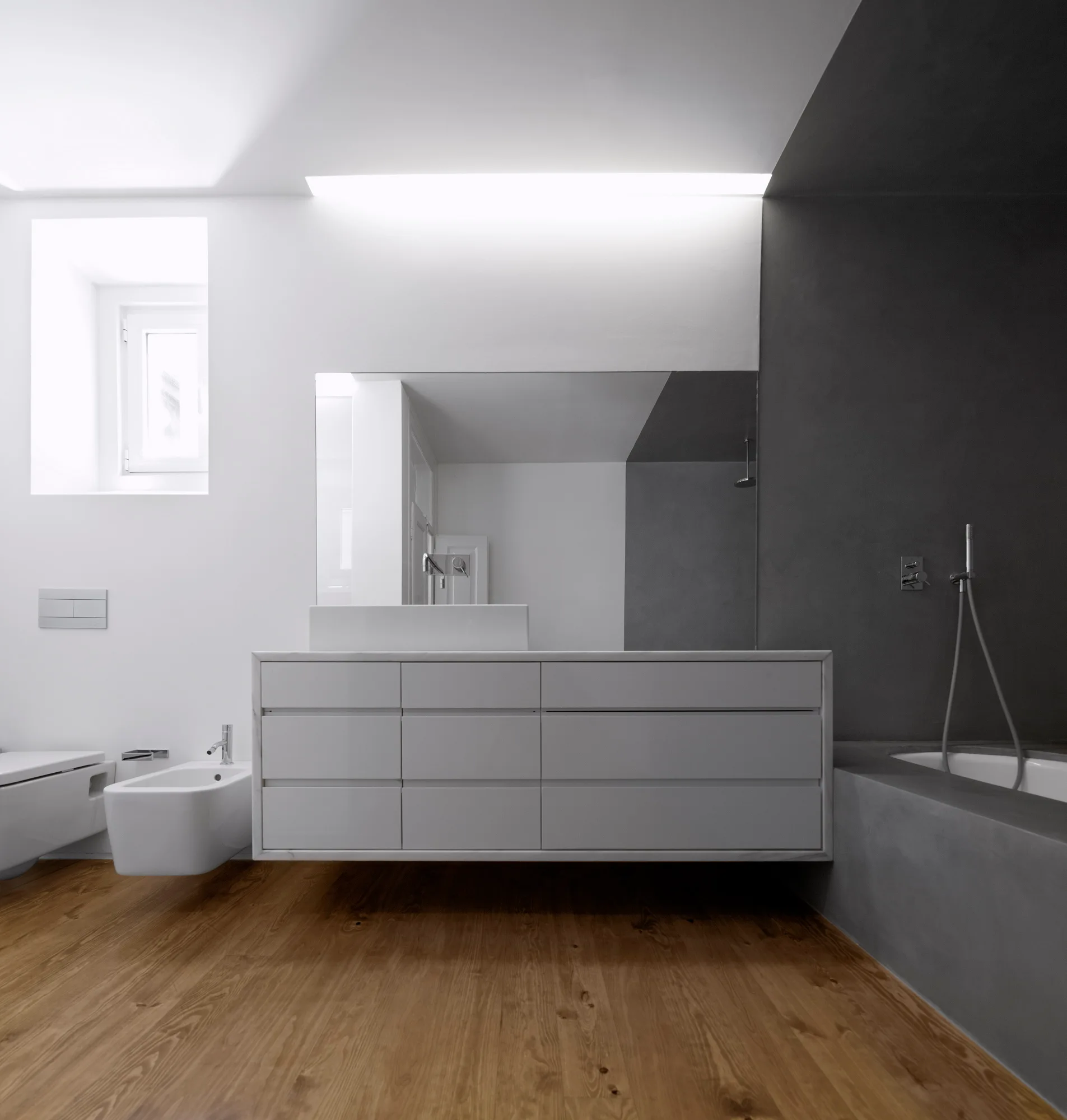 A modern minimalist bathroom features a long white floating vanity, a dark gray plaster wall with a bathtub, and warm wood plank flooring.