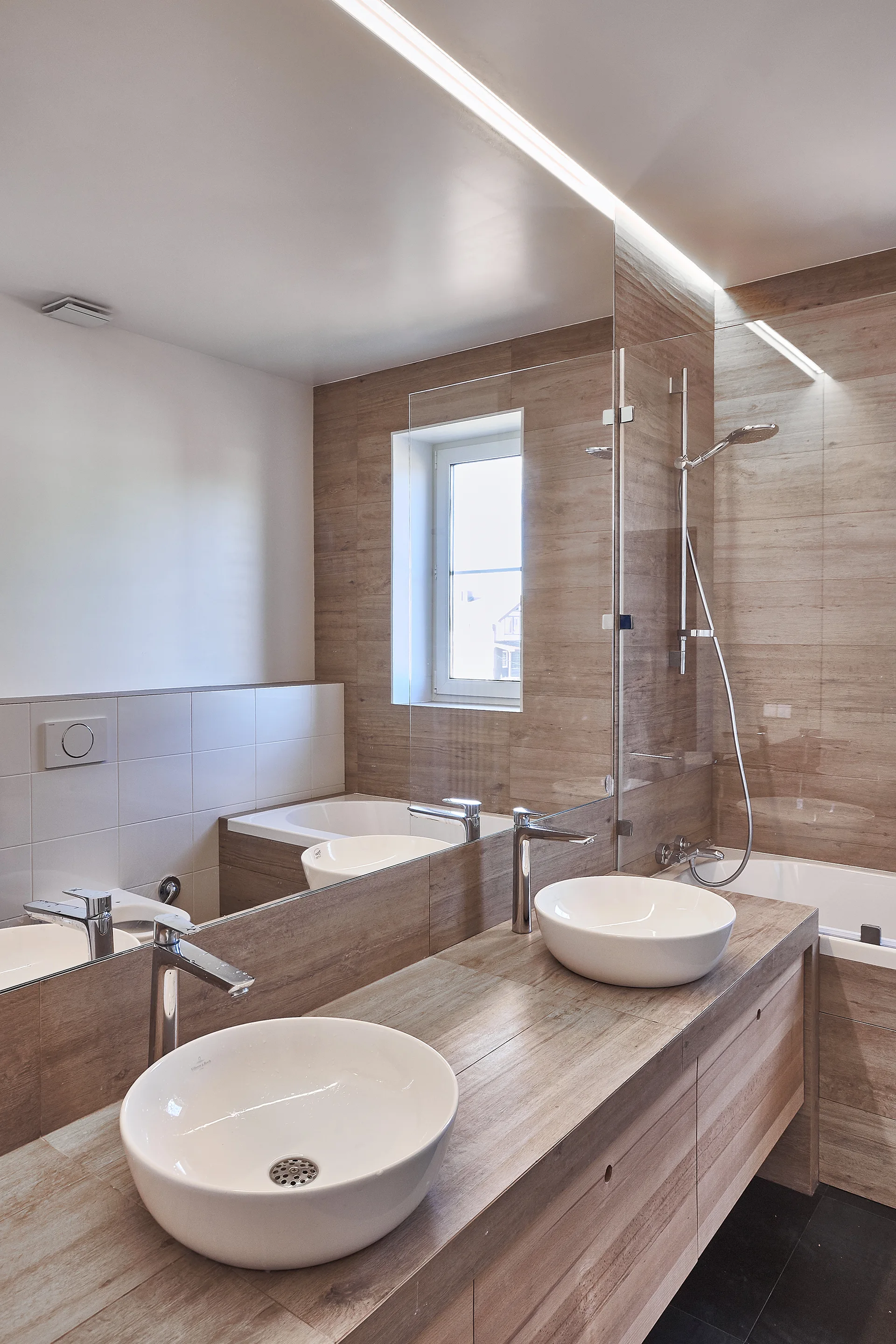A modern bathroom featuring a light wood-toned tiled wall and double vanity with two round white vessel sinks under a large wall-spanning mirror.