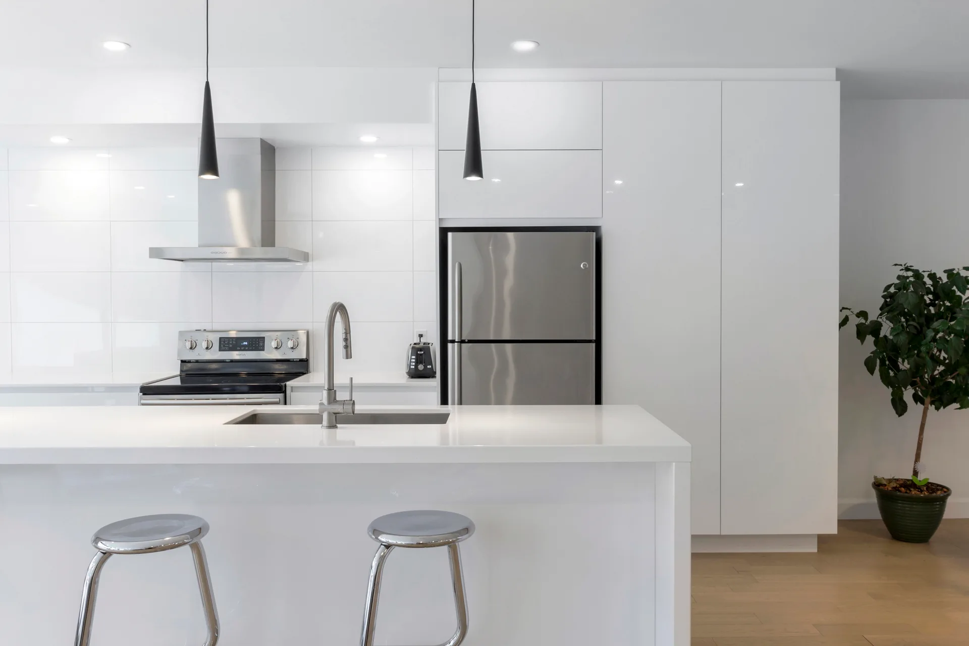A modern white kitchen with high-gloss cabinets, stainless steel appliances, a white island with two metallic bar stools, and black pendant lights.