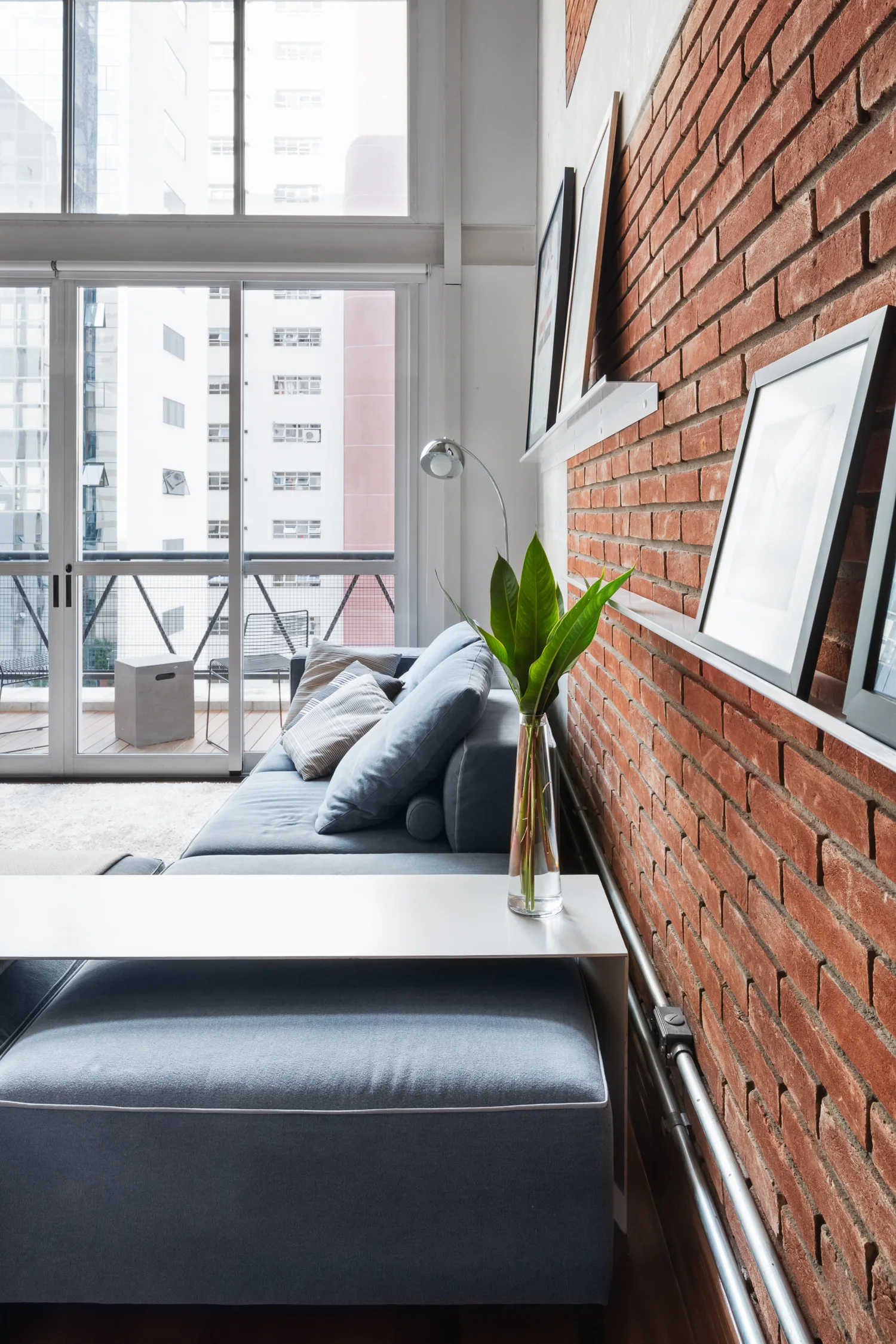 A modern loft living room features a blue modular sofa against an exposed red brick wall with white picture ledges and large windows.