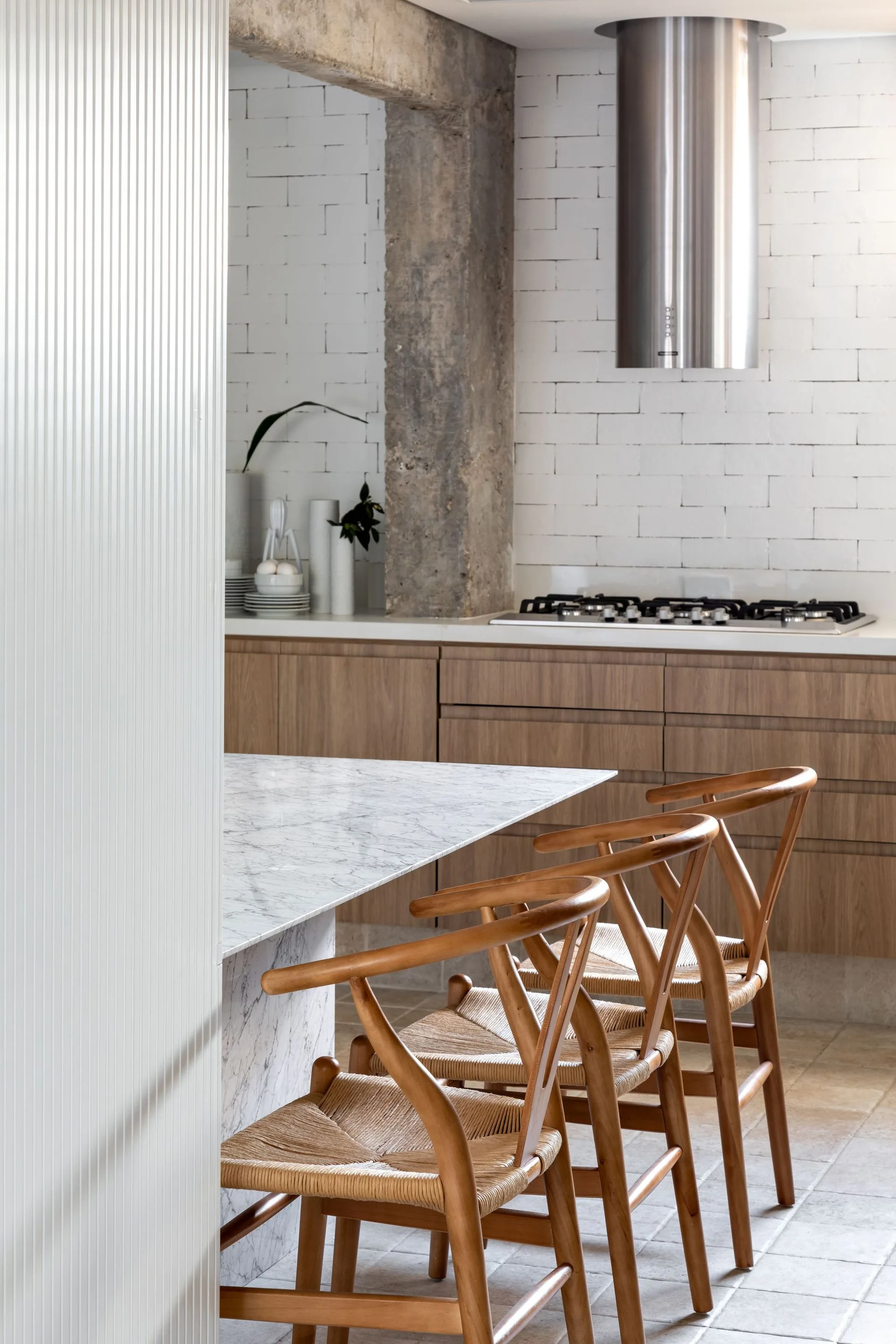 A modern kitchen featuring a marble-topped island with three wood and woven-seat stools next to an exposed concrete column and white tiled walls.