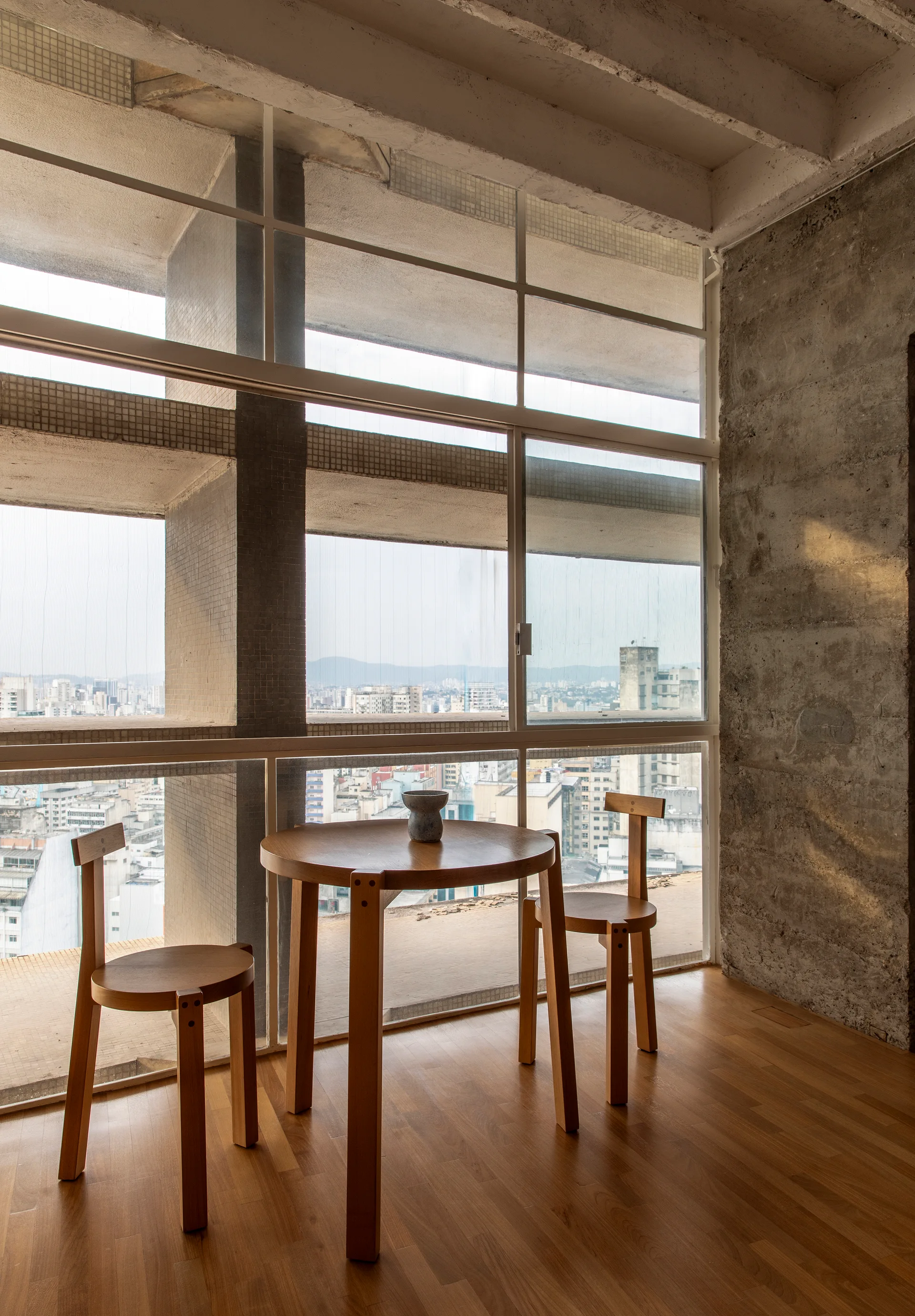 A small round wooden dining table and two matching chairs stand on a wood floor next to a large grid window and exposed concrete wall.