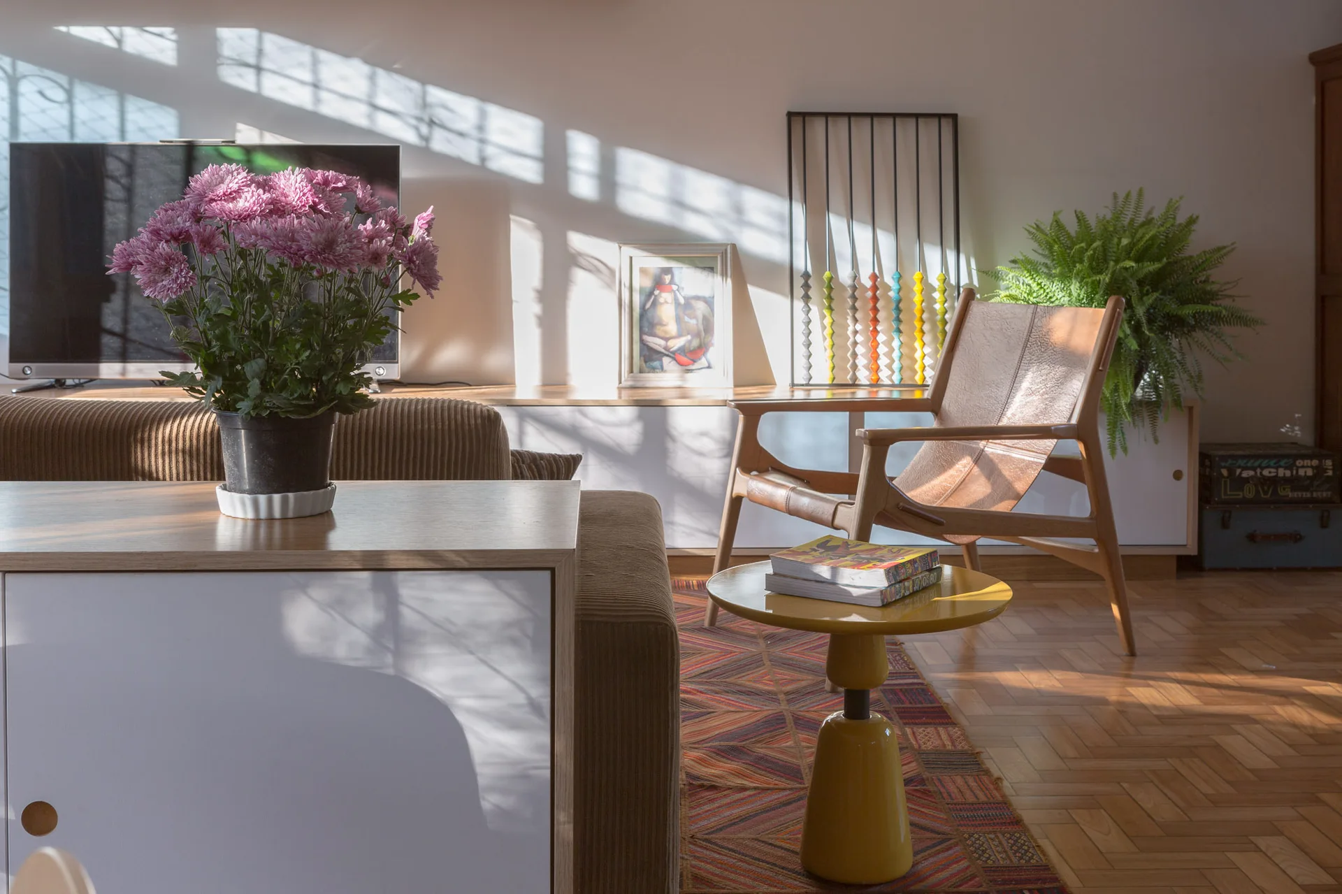 A sunlit living room featuring a leather sling chair, a yellow pedestal side table, a corduroy sofa, and shadows cast across a white wall.