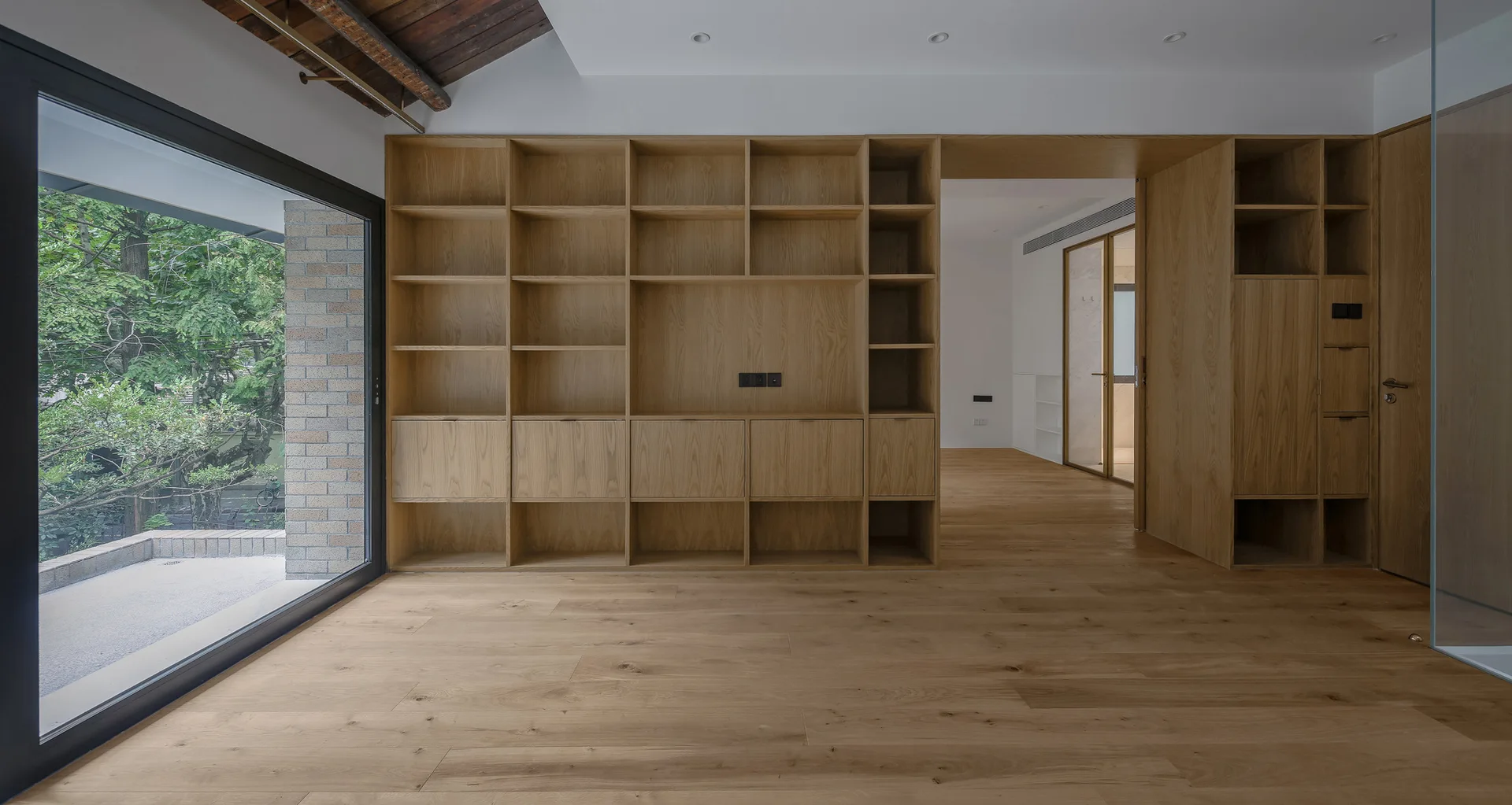 An empty minimalist room features a large custom light wood shelving unit on a white wall, matching wood floors, and a floor-to-ceiling glass door.