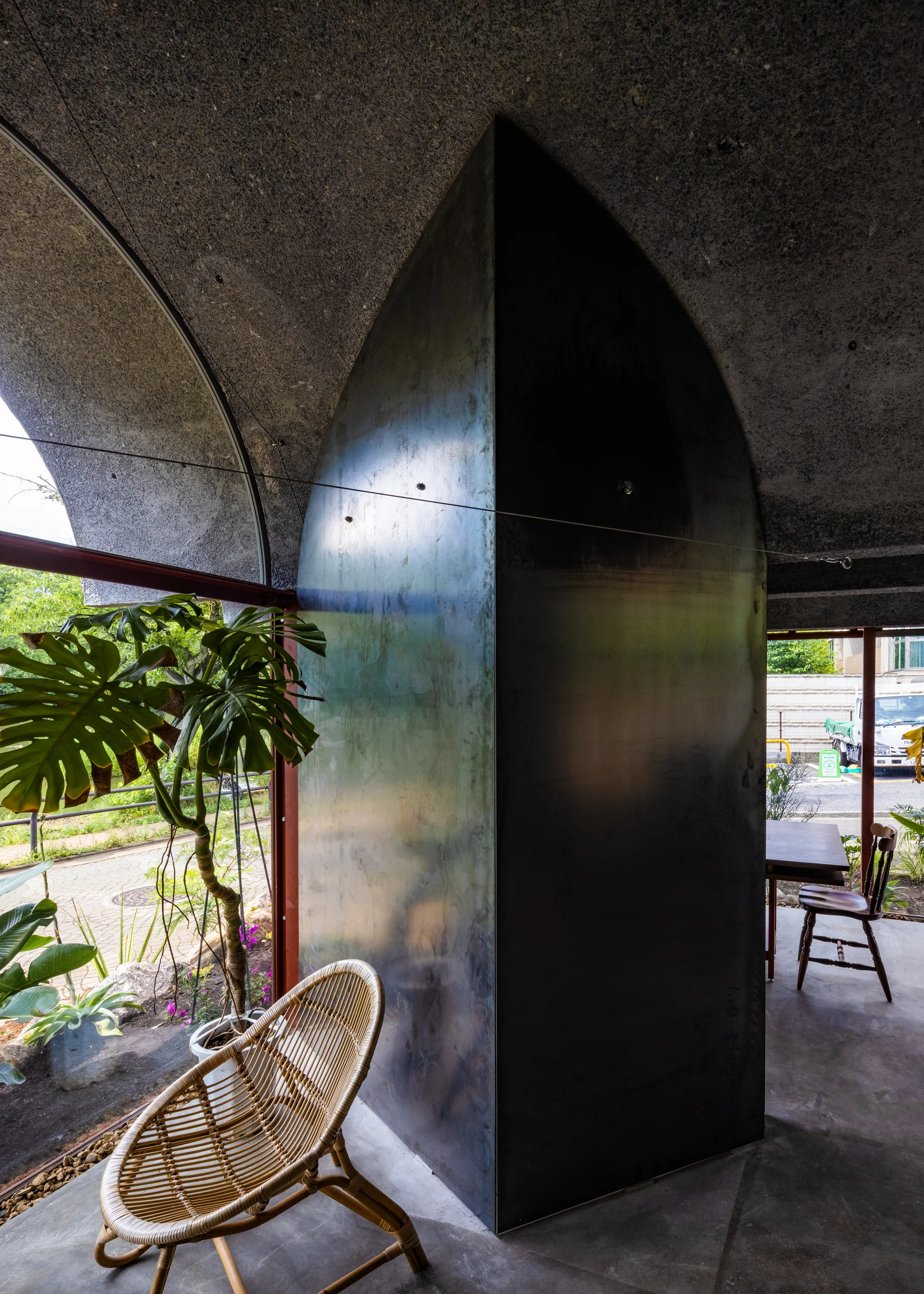 A modern interior features a textured concrete arched ceiling, a dark metallic partition wall, a rattan chair, and large windows looking outside.