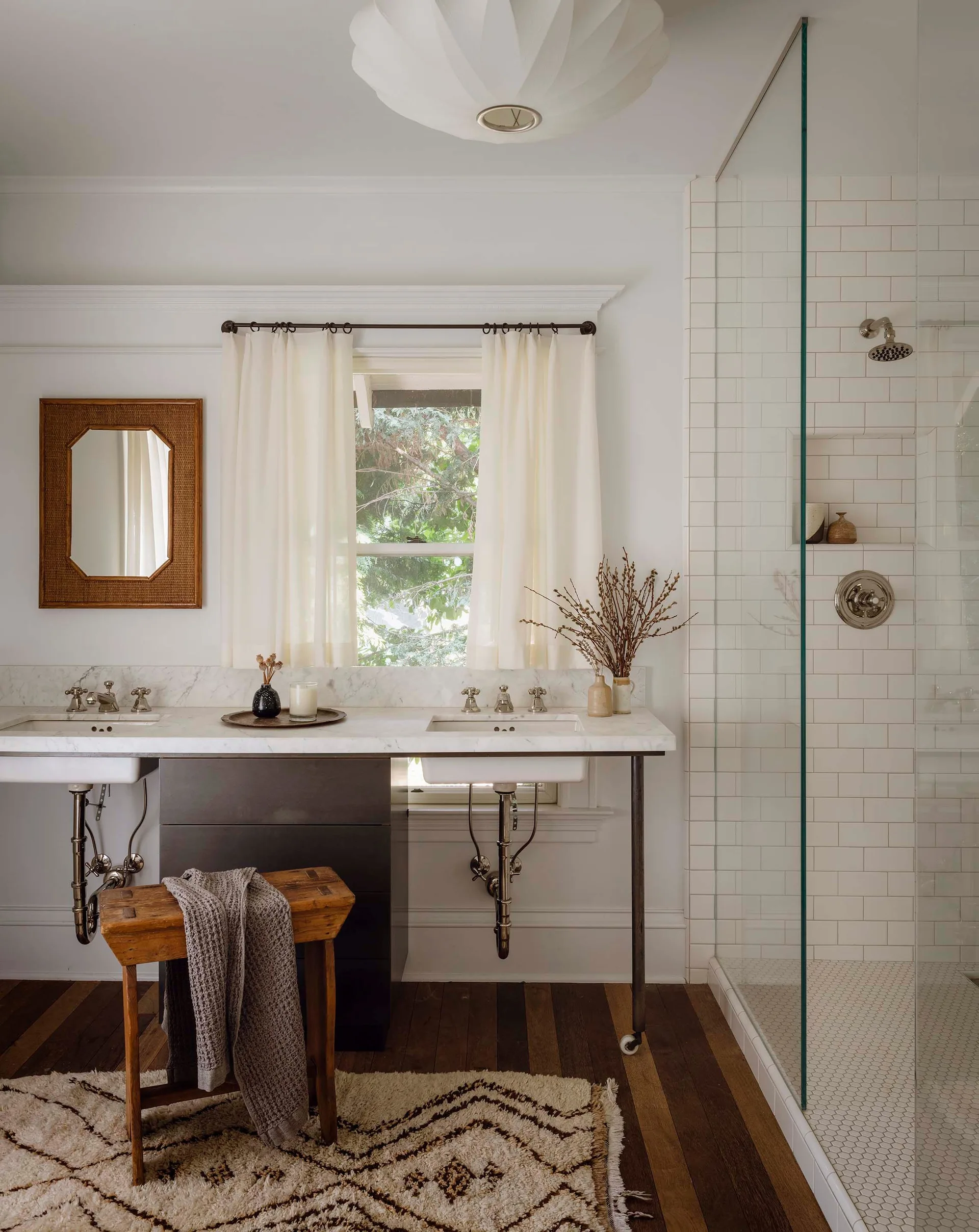 A modern bathroom features a double console vanity with exposed plumbing, a glass shower with subway tile, and a vintage wooden stool.