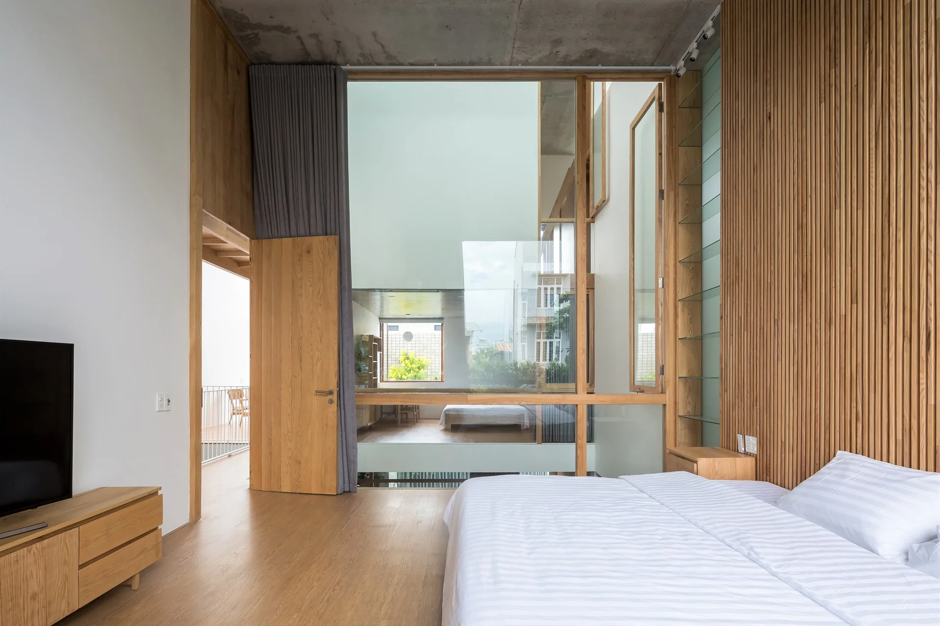 A contemporary bedroom featuring a bed with white striped bedding, a vertical wood slat accent wall, an exposed concrete ceiling, and a large glass partition.