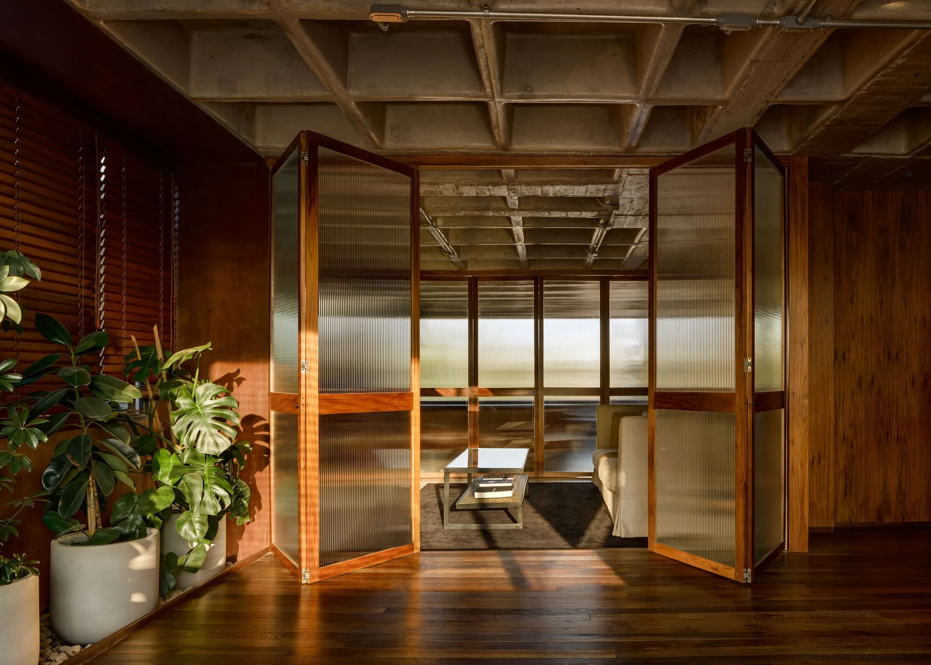 A living space features an exposed concrete waffle ceiling, warm wood-paneled walls, and open ribbed glass folding doors leading to a seating area.