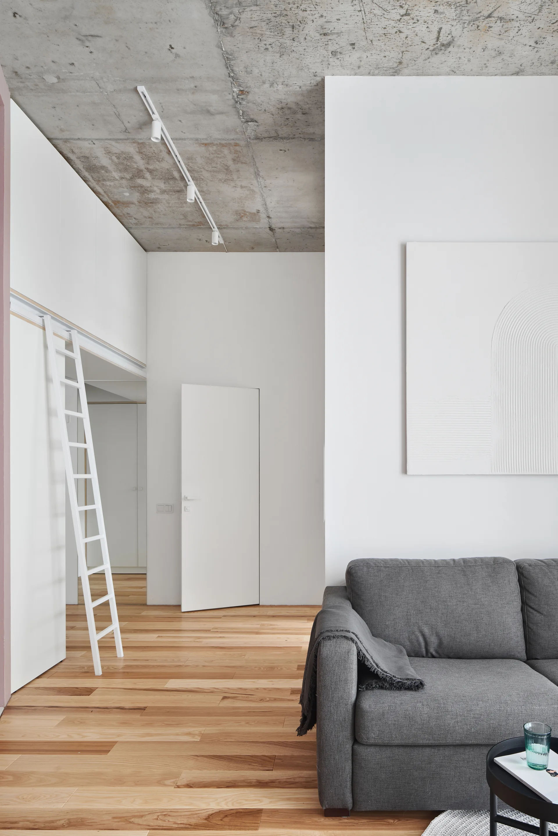 A grey sofa sits in a minimalist room with light wood floors, white built-in storage, a white ladder, and an exposed concrete ceiling.