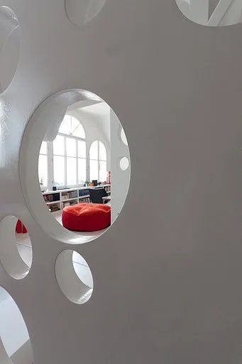 A view through a circular cutout in a white room divider reveals a bright living room with a red ottoman and arched windows.