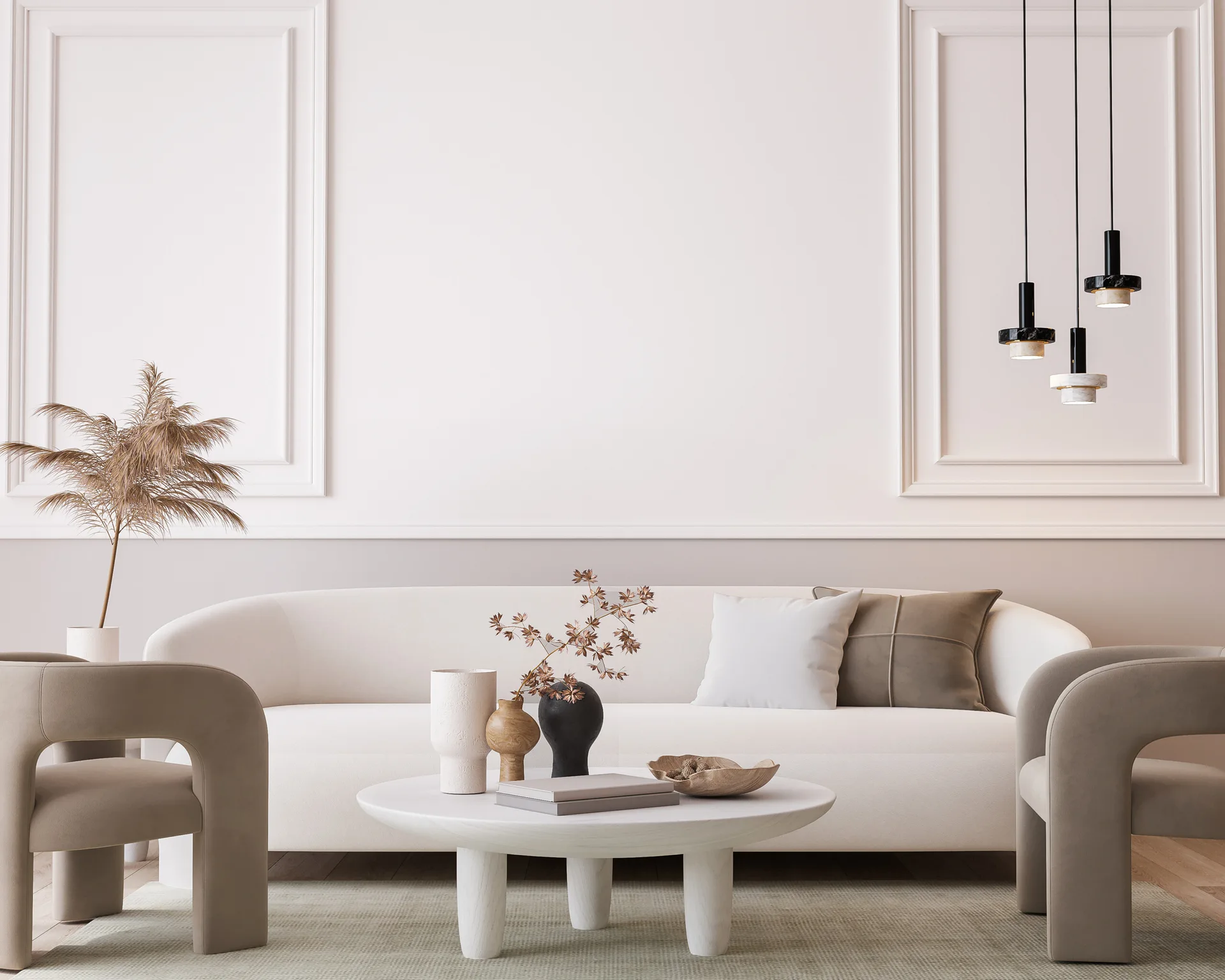 A neutral-toned living room features a white curved sofa, taupe armchairs, a round coffee table, and classical wall molding under black pendant lights.