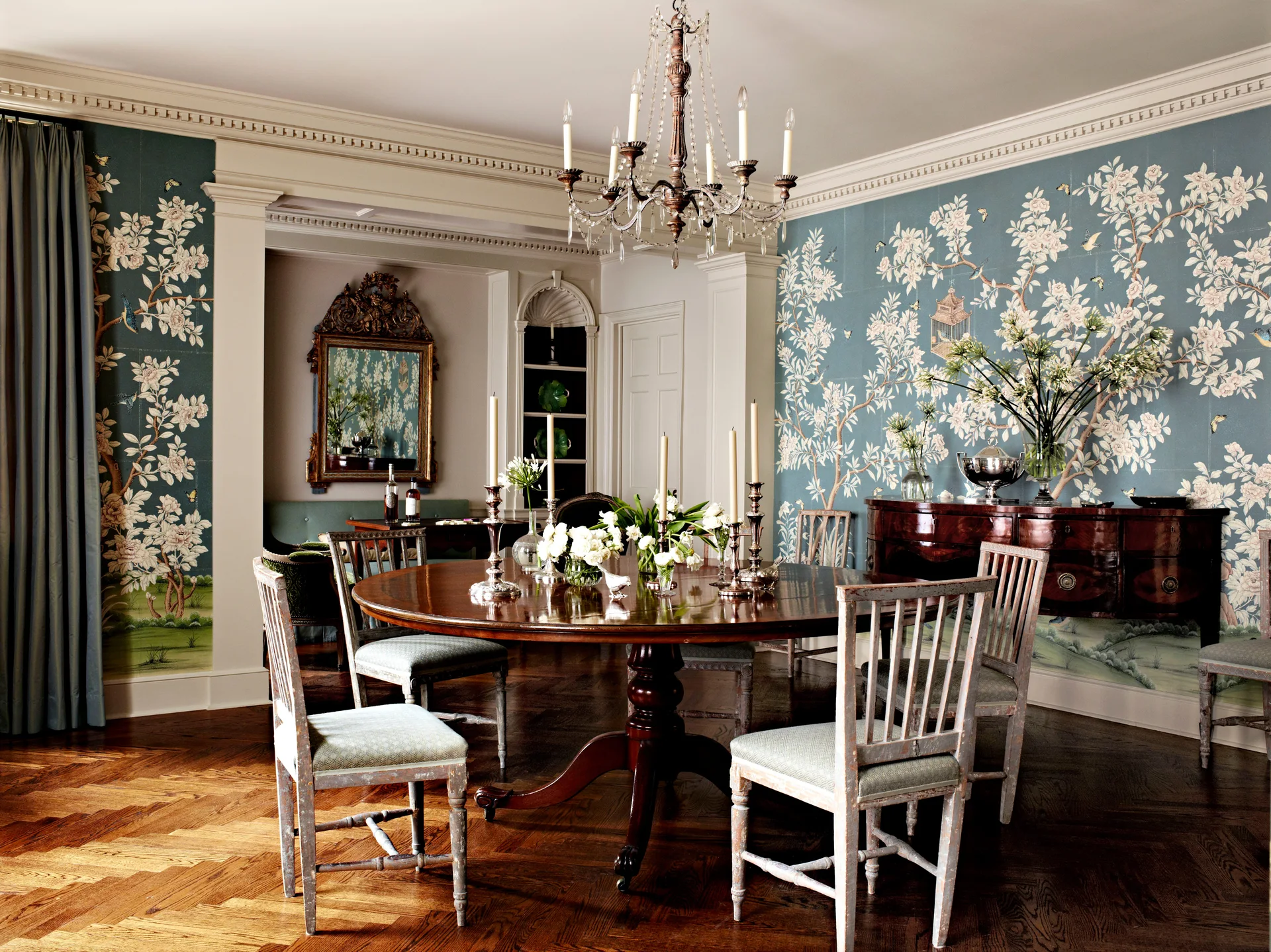 A traditional dining room features a round wooden pedestal table, distressed chairs, a crystal chandelier, and blue floral mural wallpaper.