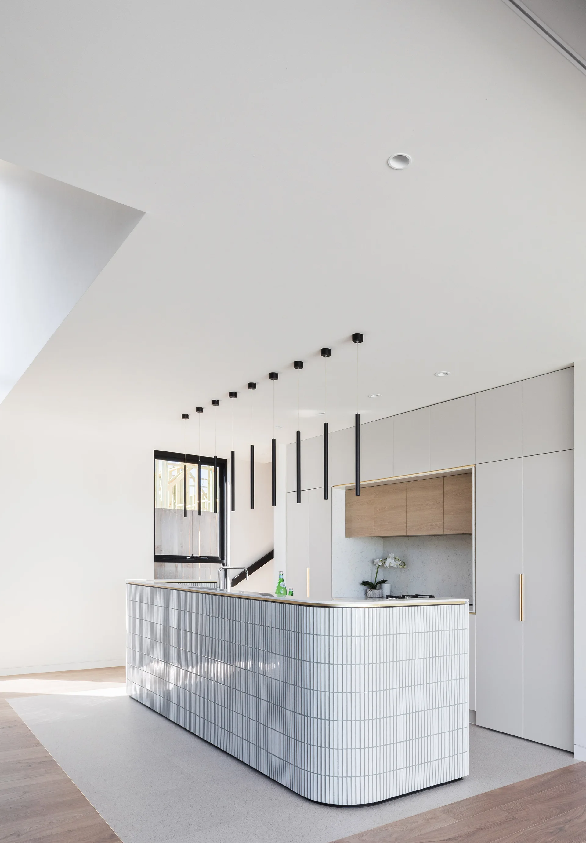 A modern kitchen features a curved island wrapped in white fluted tiles, topped with a row of slender black cylinder pendant lights.